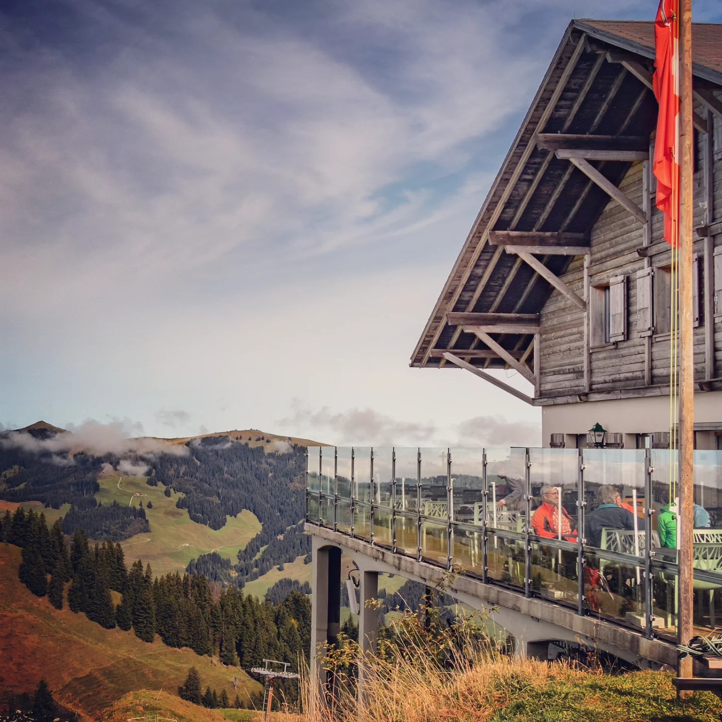 Wooden mountain restaurant with a glass-railed terrace cantilevered over a slope, diners in colorful jackets inside.