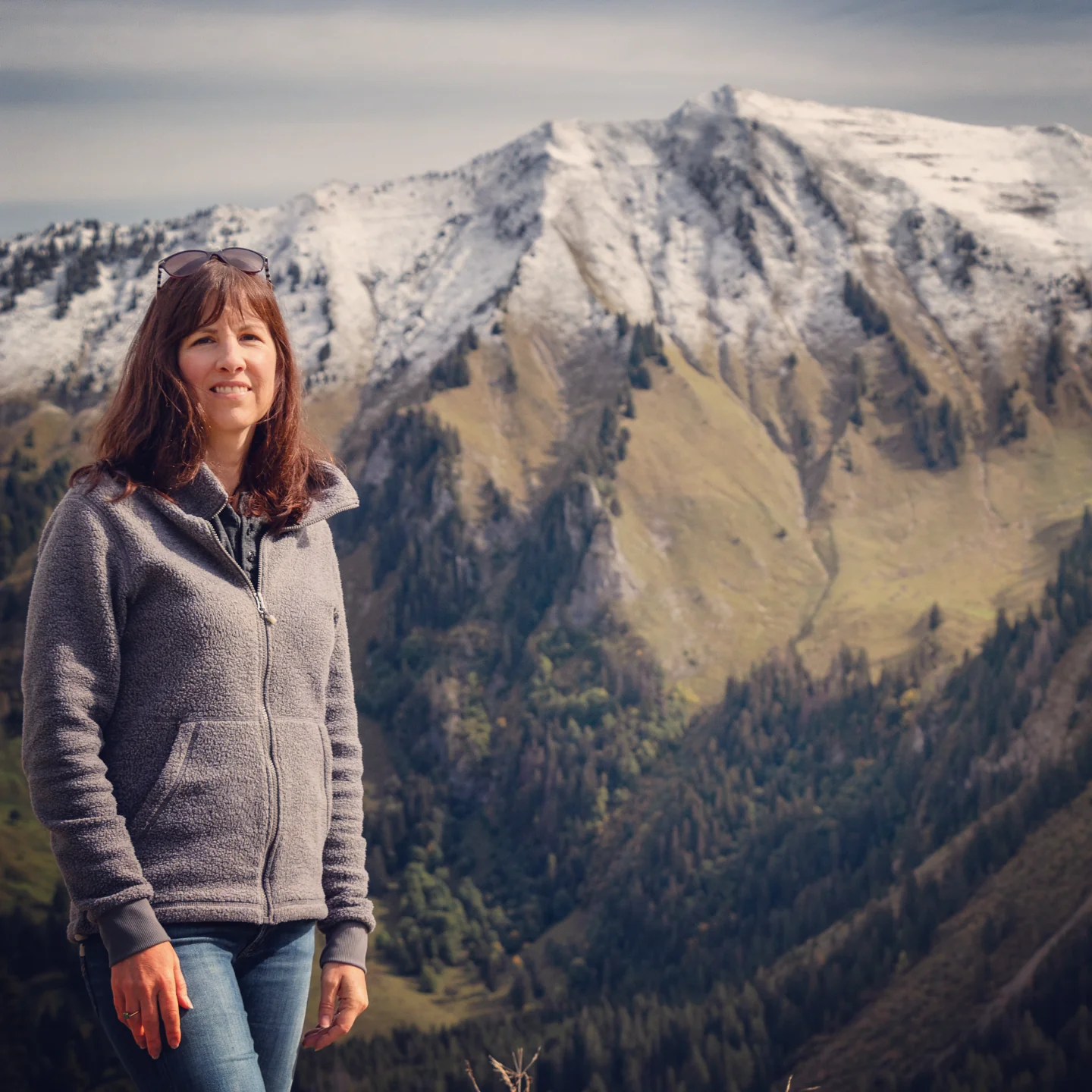 Woman in grey fleece poses on a ridge; behind her a freshly snow-dusted peak rises above forested slopes.
