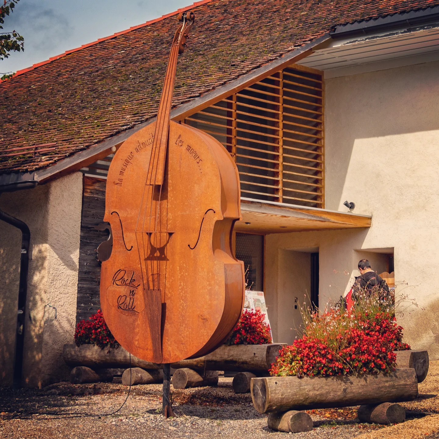 Weathered steel double-bass sculpture with the French inscription “La musique adoucit les mœurs,” standing outside a low stucco building in the Swiss hill-town of Gruyères.