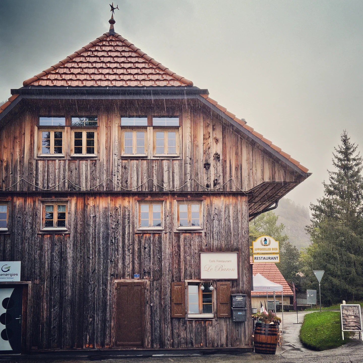 A rain-soaked, three-storey timber chalet with a red-tiled roof and the sign “Café Restaurant Le Baron” on its weathered façade in Charmey, Switzerland.