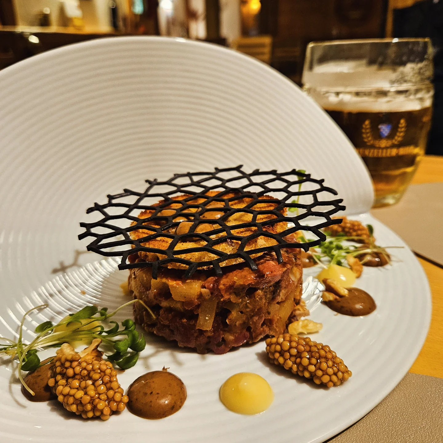 Close-up of an artfully plated beef tartare topped with a black lattice tuile, surrounded by pickled mustard seeds, sprouts and sauces; a glass of beer sits behind.