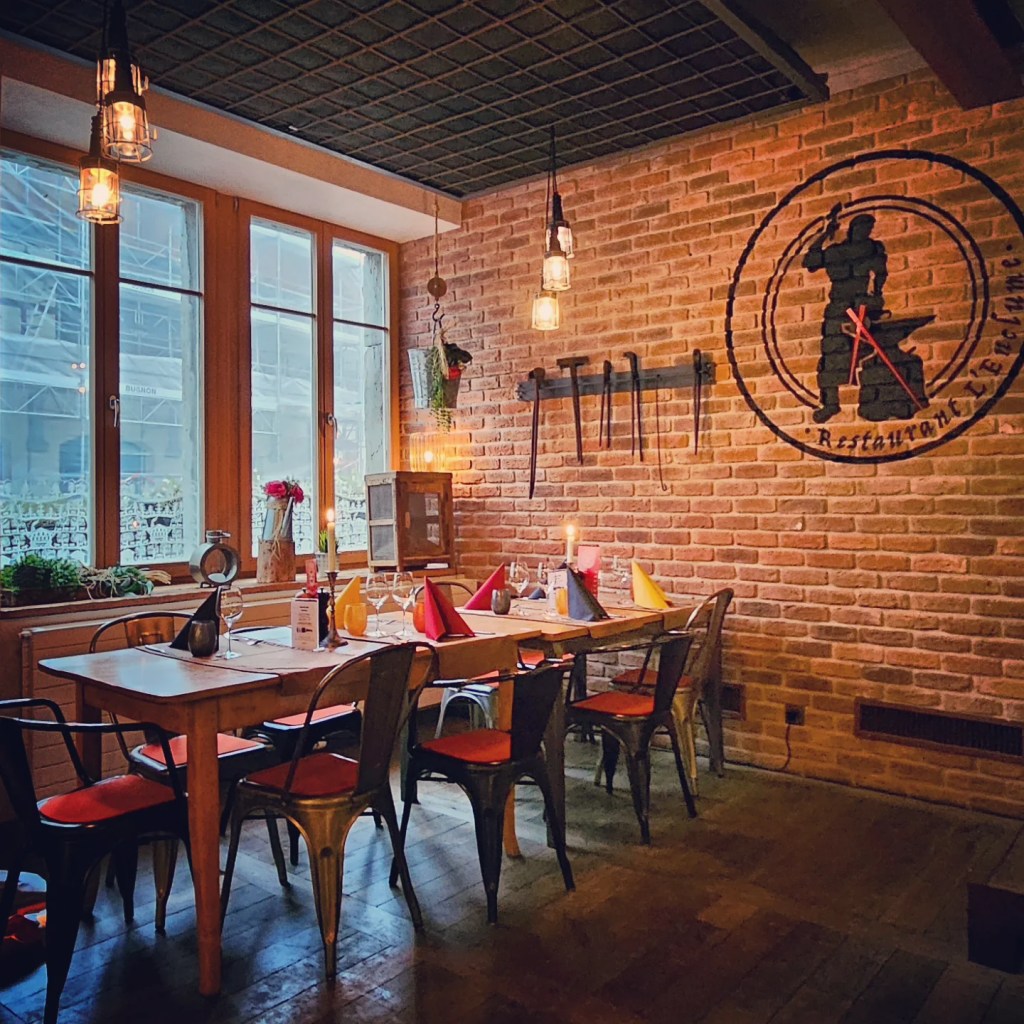 Brick-walled dining space with blacksmith tools mounted on the wall, a long wooden table set with multicolored napkins and hanging filament bulbs.