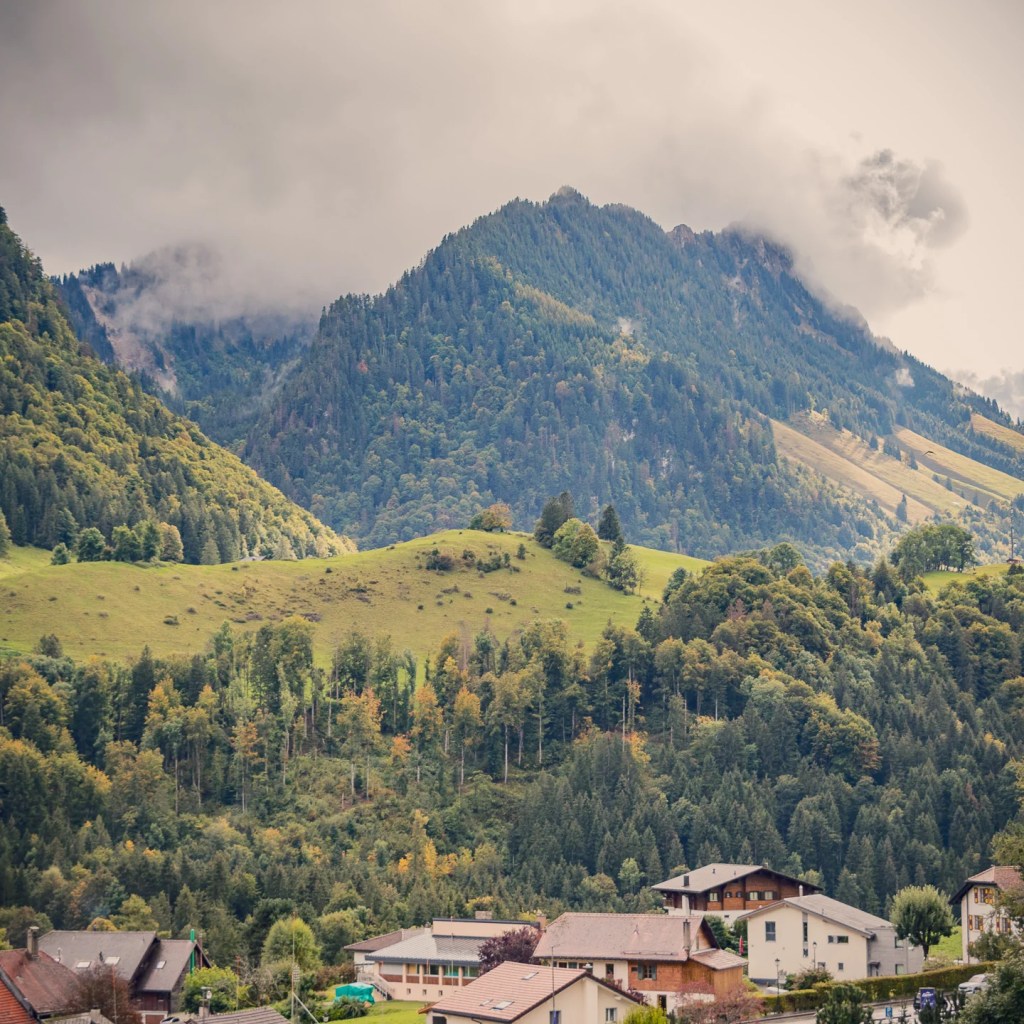 View from a hotel terrace in Charmey, Switzerland—village rooftops in the foreground, green pastures and mixed forest in the mid-ground, and a cloud-capped Pre-Alps peak rising behind.