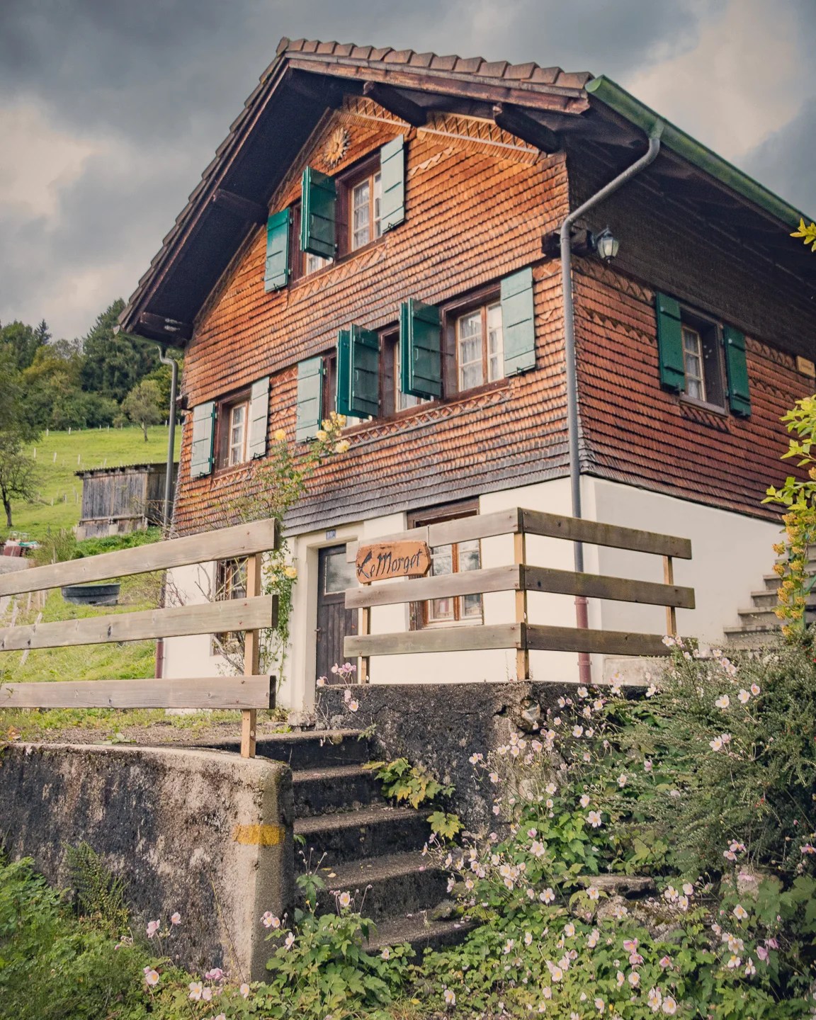 Timber-shingled alpine cottage “Le Morget” with green shutters, stone steps, and wildflowers.