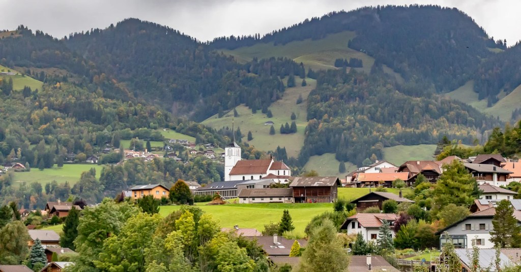 Wide view of Saint-Pierre-aux-Liens church and farm buildings on a ridge, framed by forested mountains.