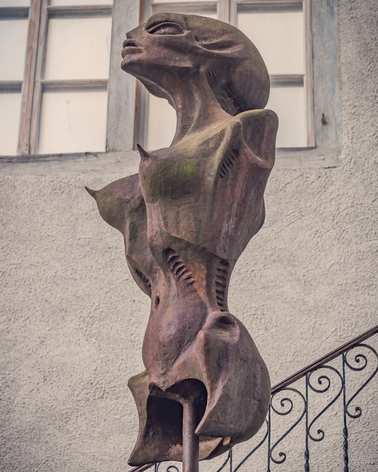 Weathered bronze biomechanical female torso sculpture by Swiss artist H. R. Giger on a pedestal beside a wrought-iron stair outside a stone building in Gruyères.