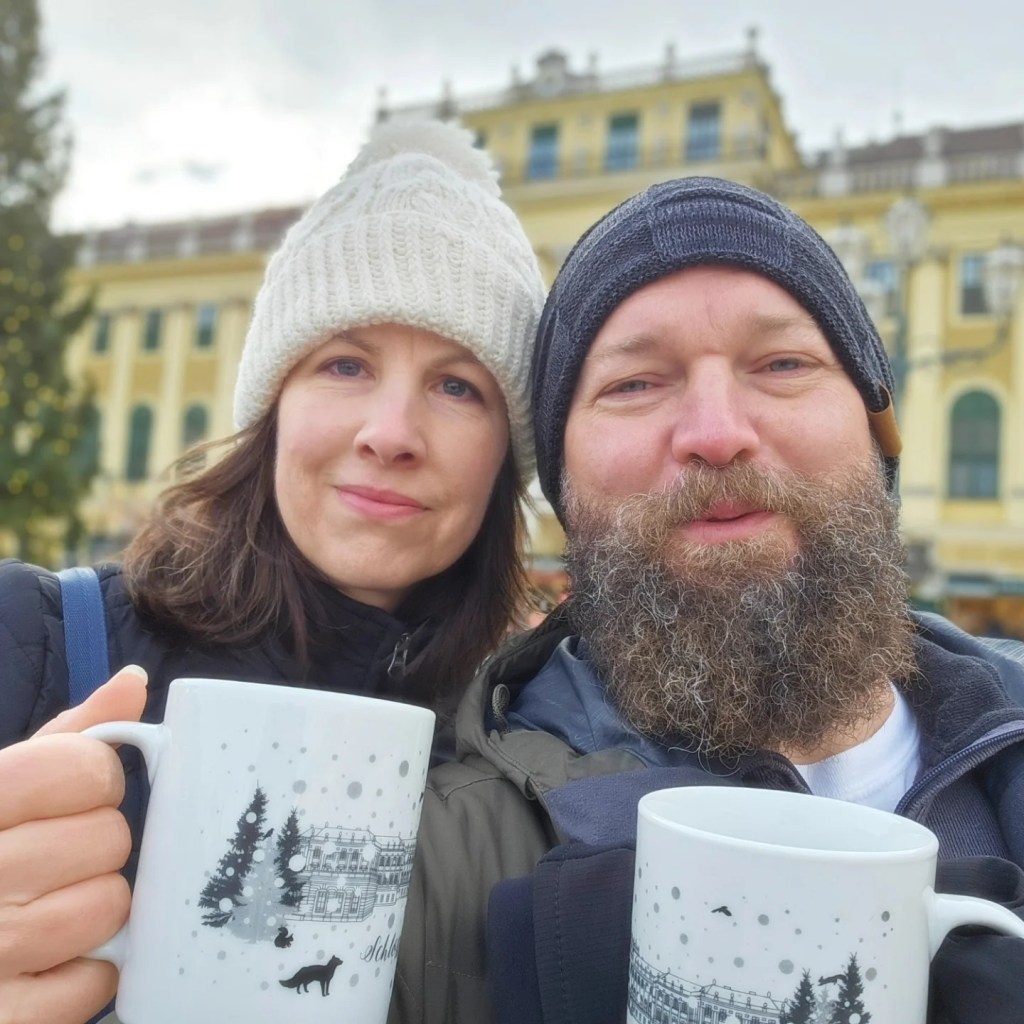 A couple bundled up in winter hats holding festive mugs at a Christmas market in Vienna, with the yellow Schönbrunn Palace in the background.