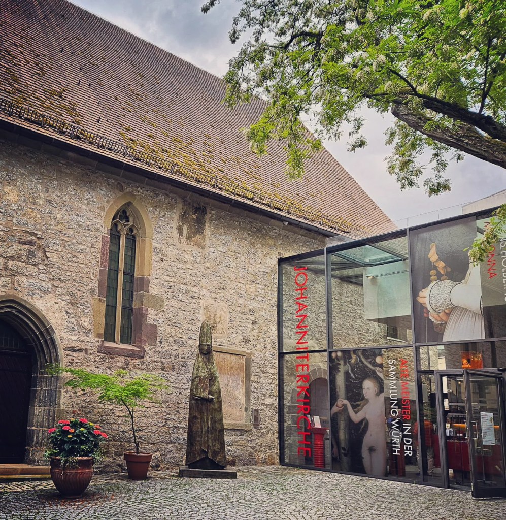 The stone exterior of the Johanniterkirche Museum in Schwäbisch Hall, featuring Gothic-style arched windows, a bronze bishop statue, and a modern glass entrance with large exhibition banners.