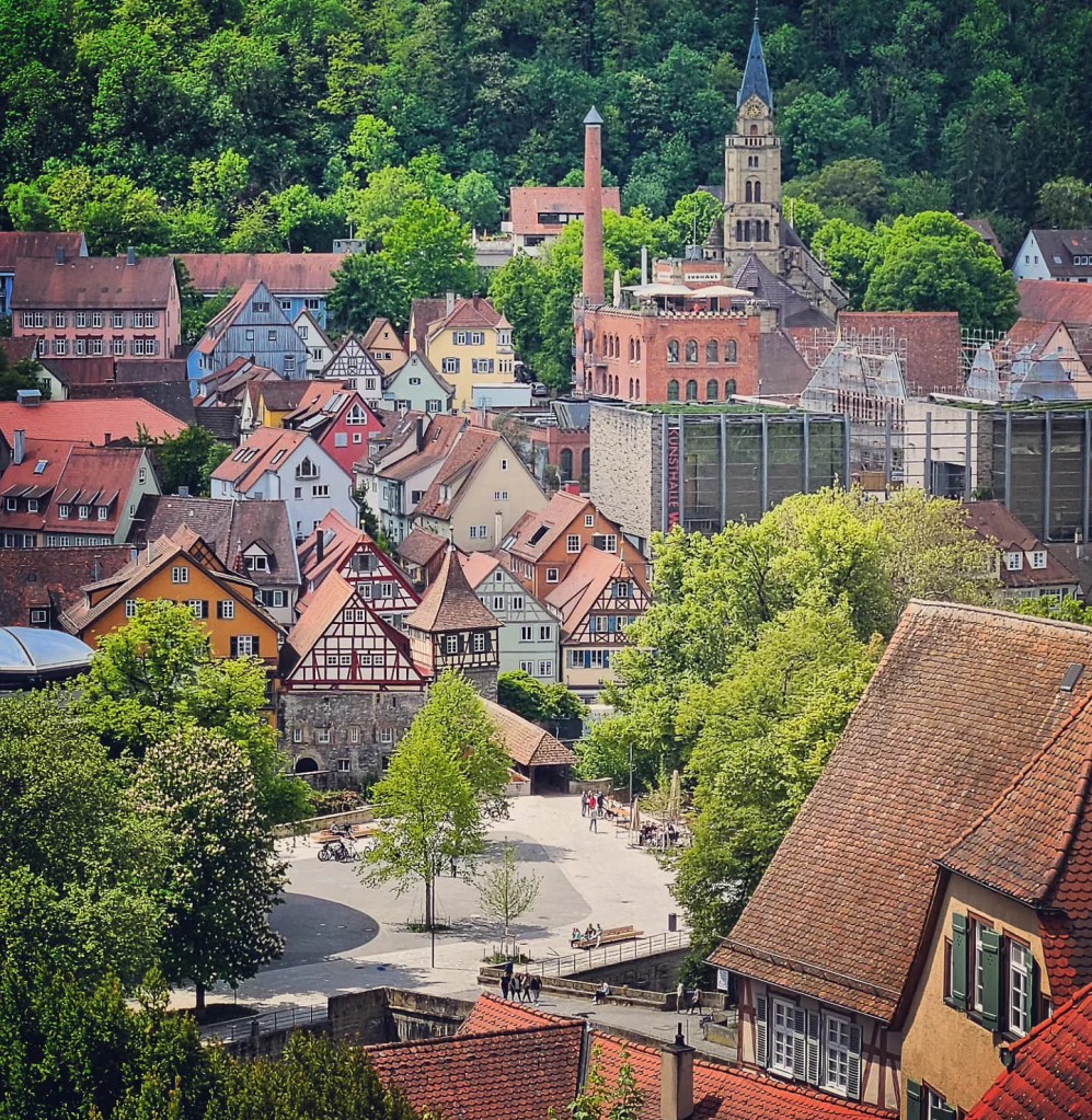 A panoramic view over the colorful half-timbered houses and red-tiled rooftops of Schwäbisch Hall, framed by green hills and with the tower of St. Katharina Church visible in the background.