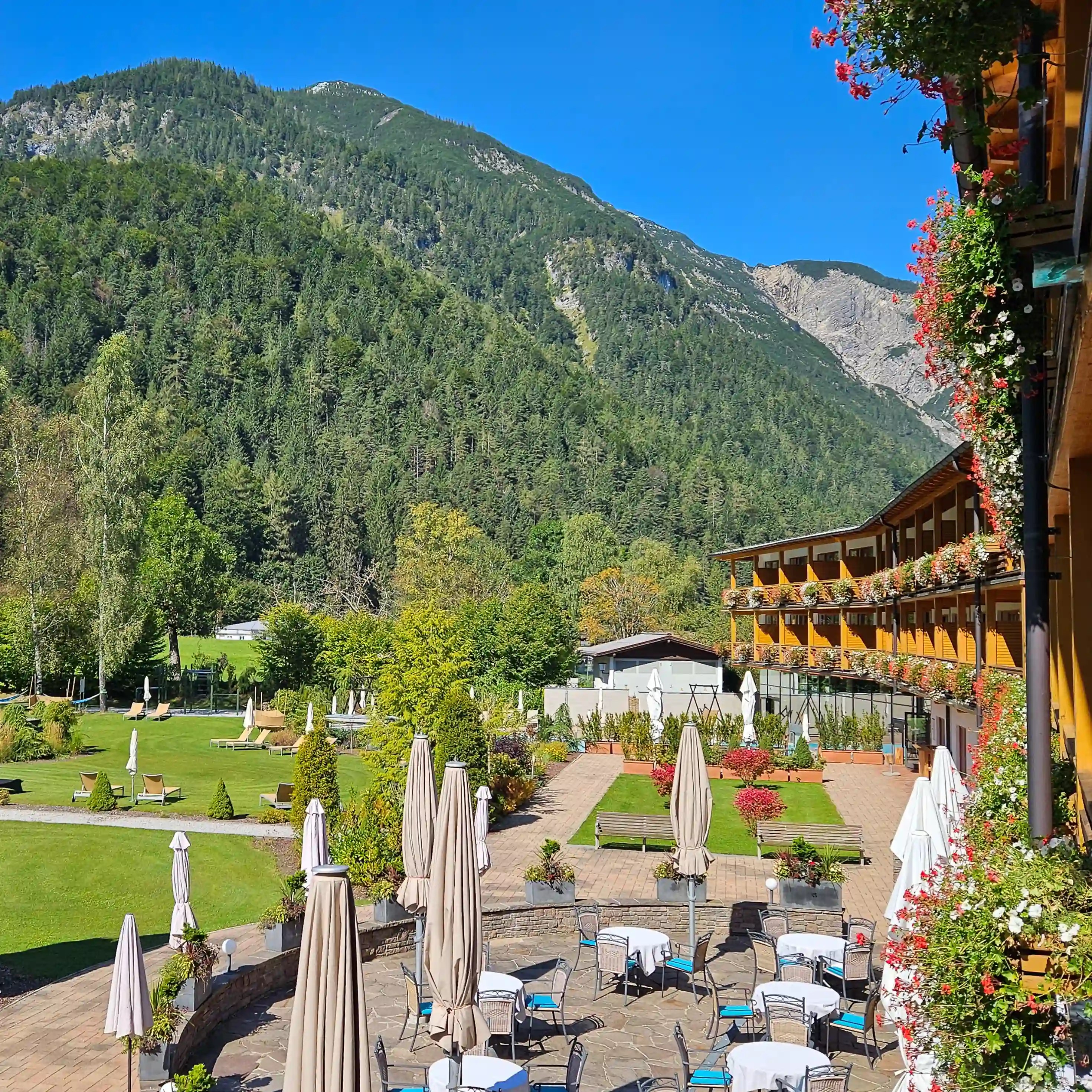 Terrace and gardens of a lakeside hotel in Pertisau with flower-decked balconies, cafe tables, and forested mountains under a bright blue sky.