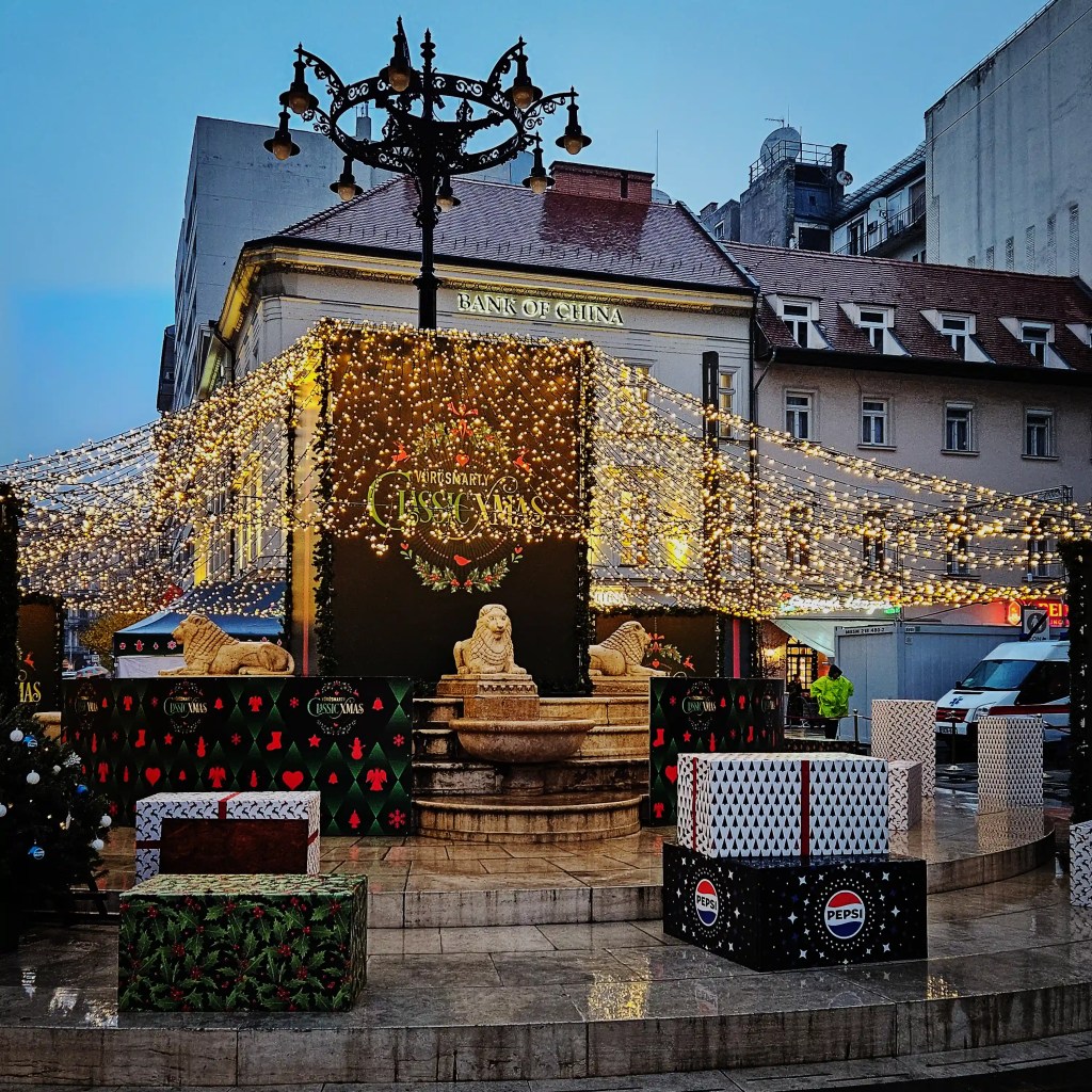 Light-covered lion fountain and gift boxes at Vörösmarty Square Christmas Market in Budapest.