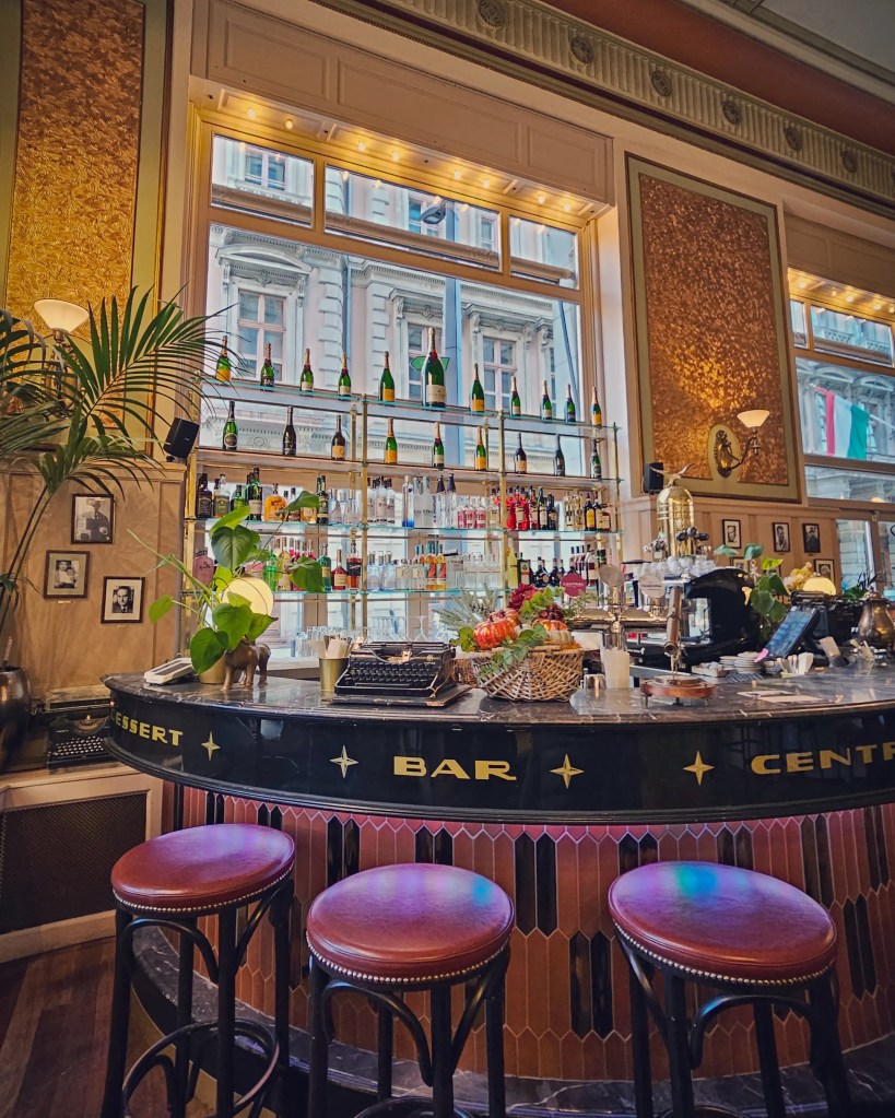 An elegant vintage bar with leather stools, champagne bottles, and art deco decor inside Budapest’s historic Central Café.