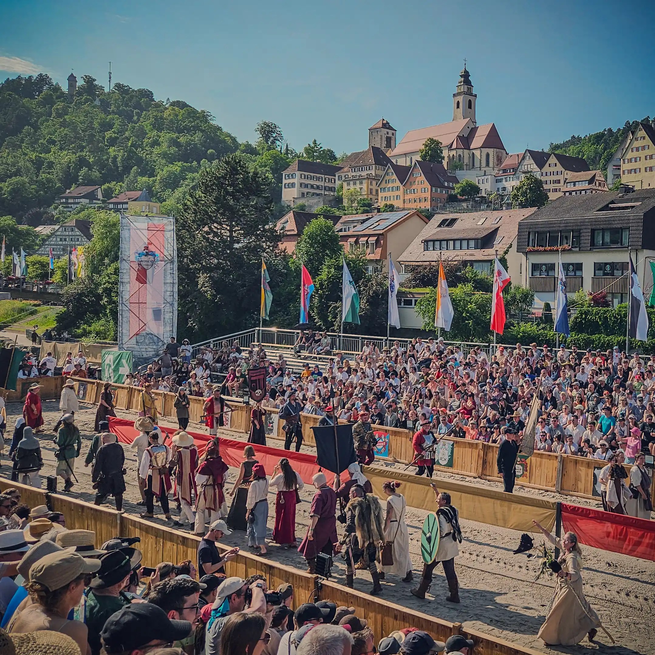 Costumed performers in medieval attire parade through an arena during the Horber Ritterspiele in Horb am Neckar, Germany, with the town’s historic buildings and church in the background.