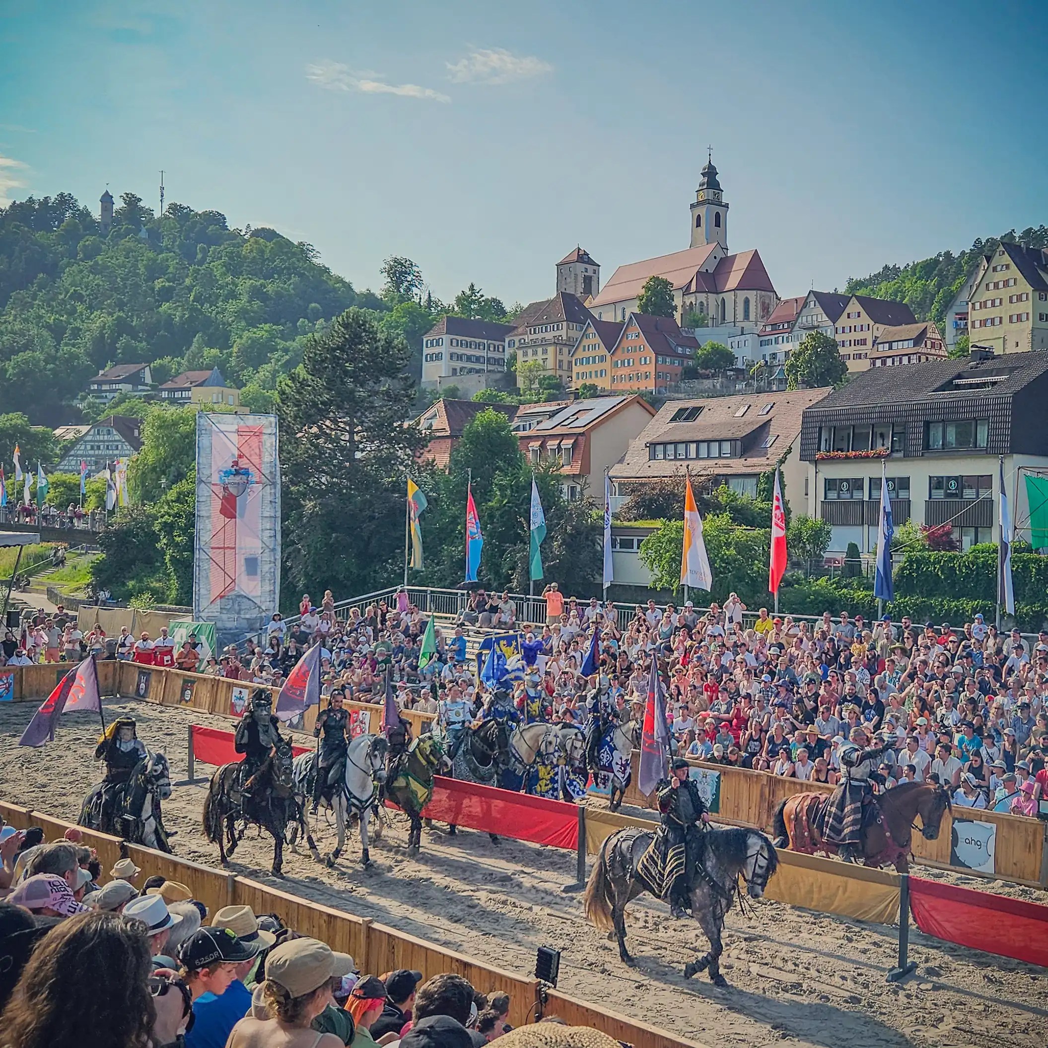 A group of costumed riders on horseback parades through the arena at the Horber Ritterspiele, with spectators lining the stands and colorful flags flying.