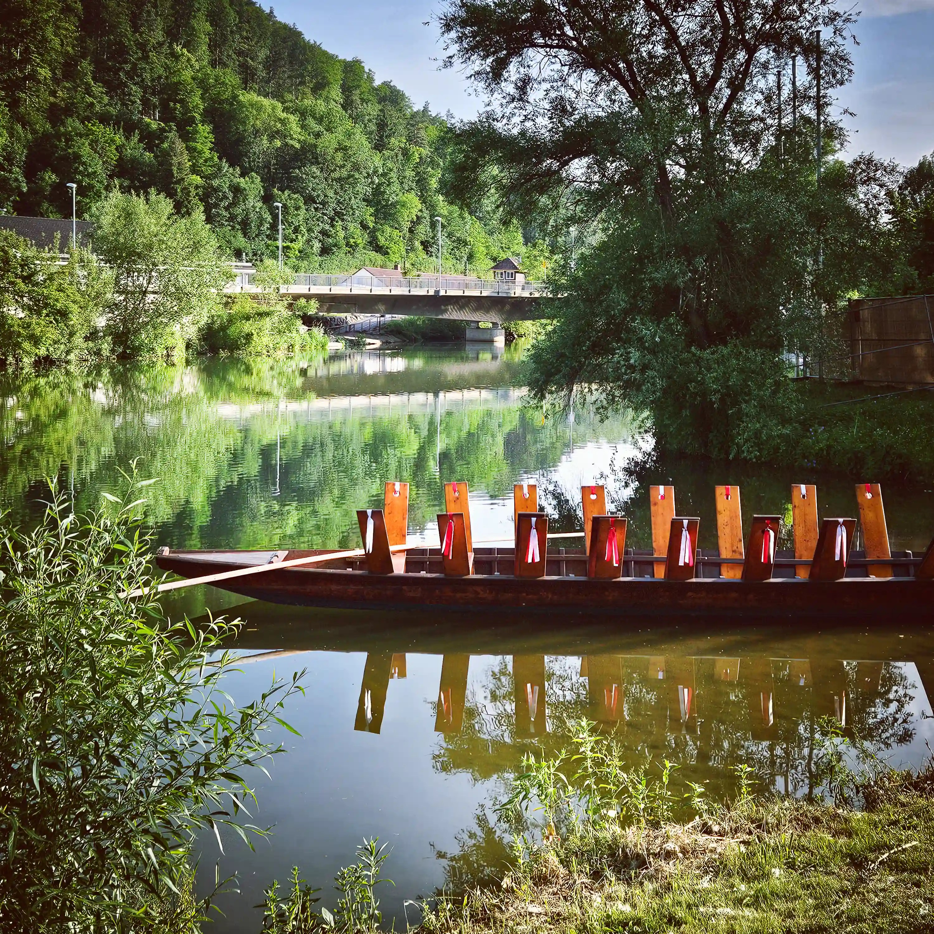 Traditional wooden boat with upright seats and red-and-white ribbons moored on the calm Neckar River in Horb am Neckar, surrounded by lush green trees and a bridge in the background.