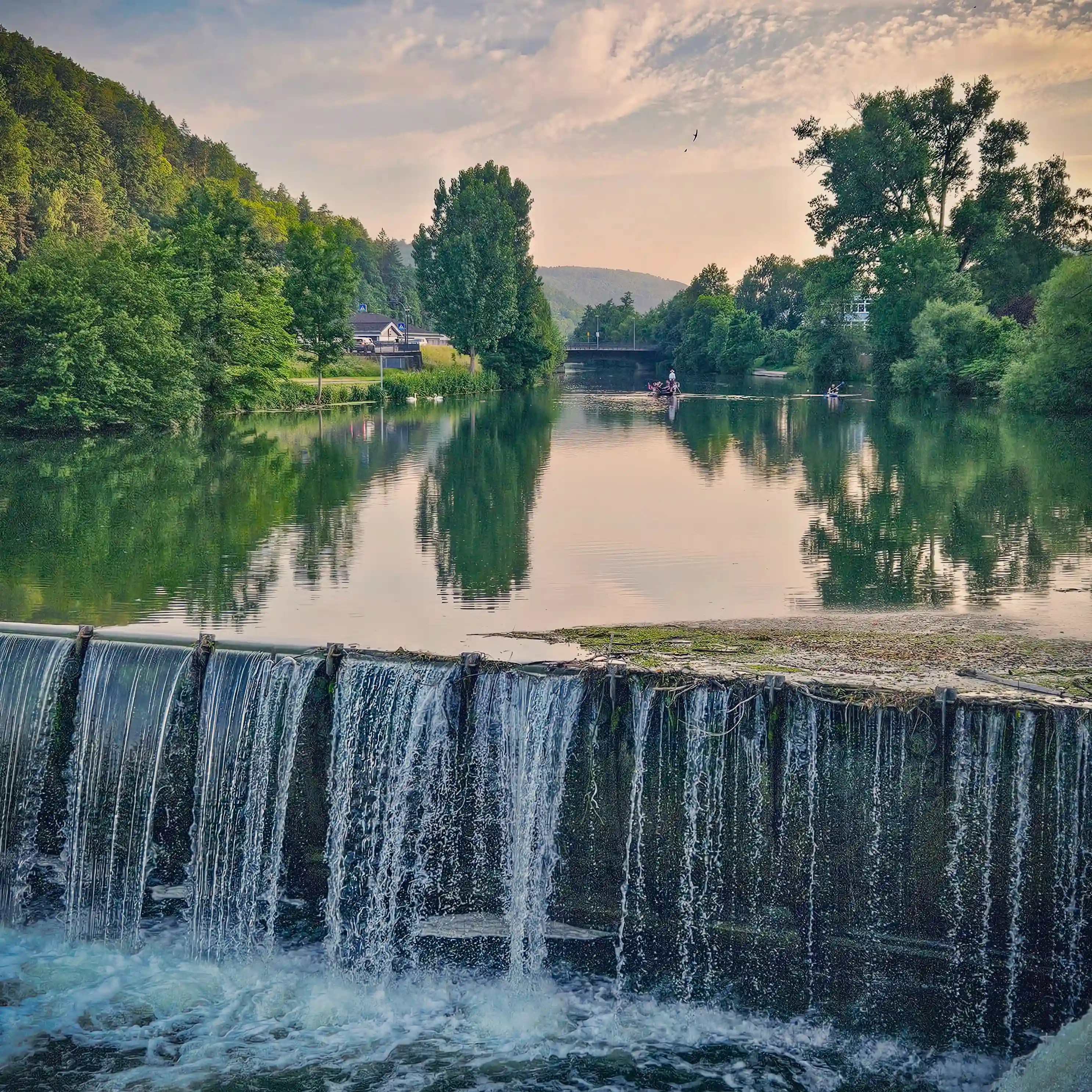 A small waterfall over a weir along the Neckar River at sunset, with trees and reflections glowing under a soft sky.