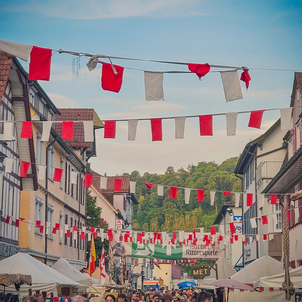 Red and white medieval-style flags hang above a busy festival street at the Horber Ritterspiele, with booths and people lining the way.