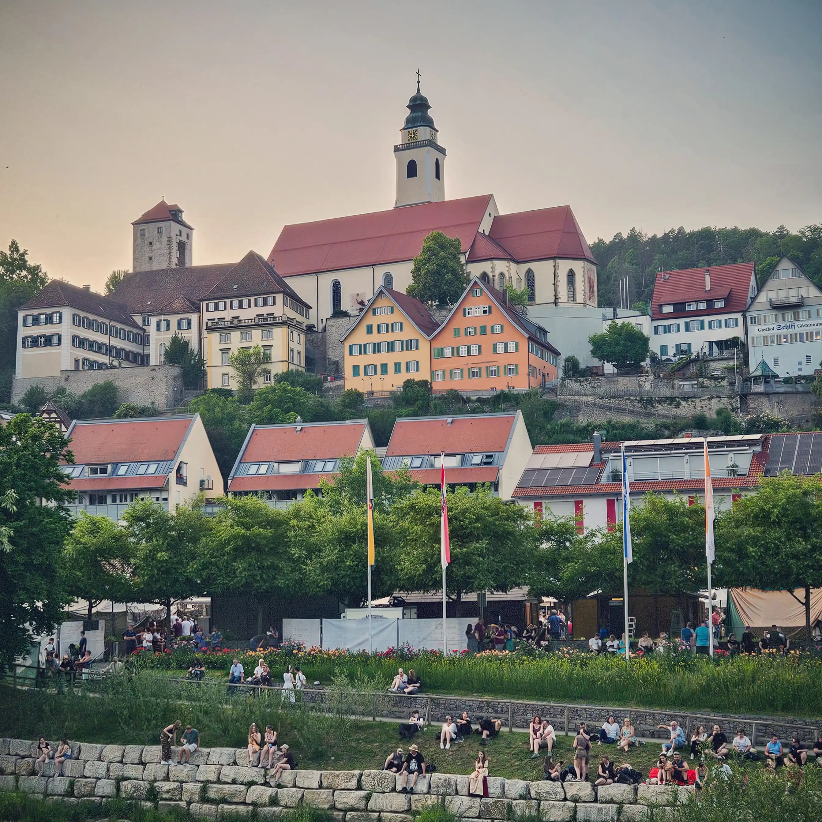 People relaxing on a grassy riverbank with a view of Horb am Neckar’s church and colorful hillside buildings in the background.