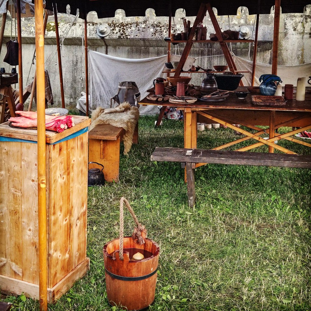 A rustic medieval camp kitchen at the Horb festival, with wooden tables, benches, clay dishes, fur-covered seating, and a wooden bucket on the grass.