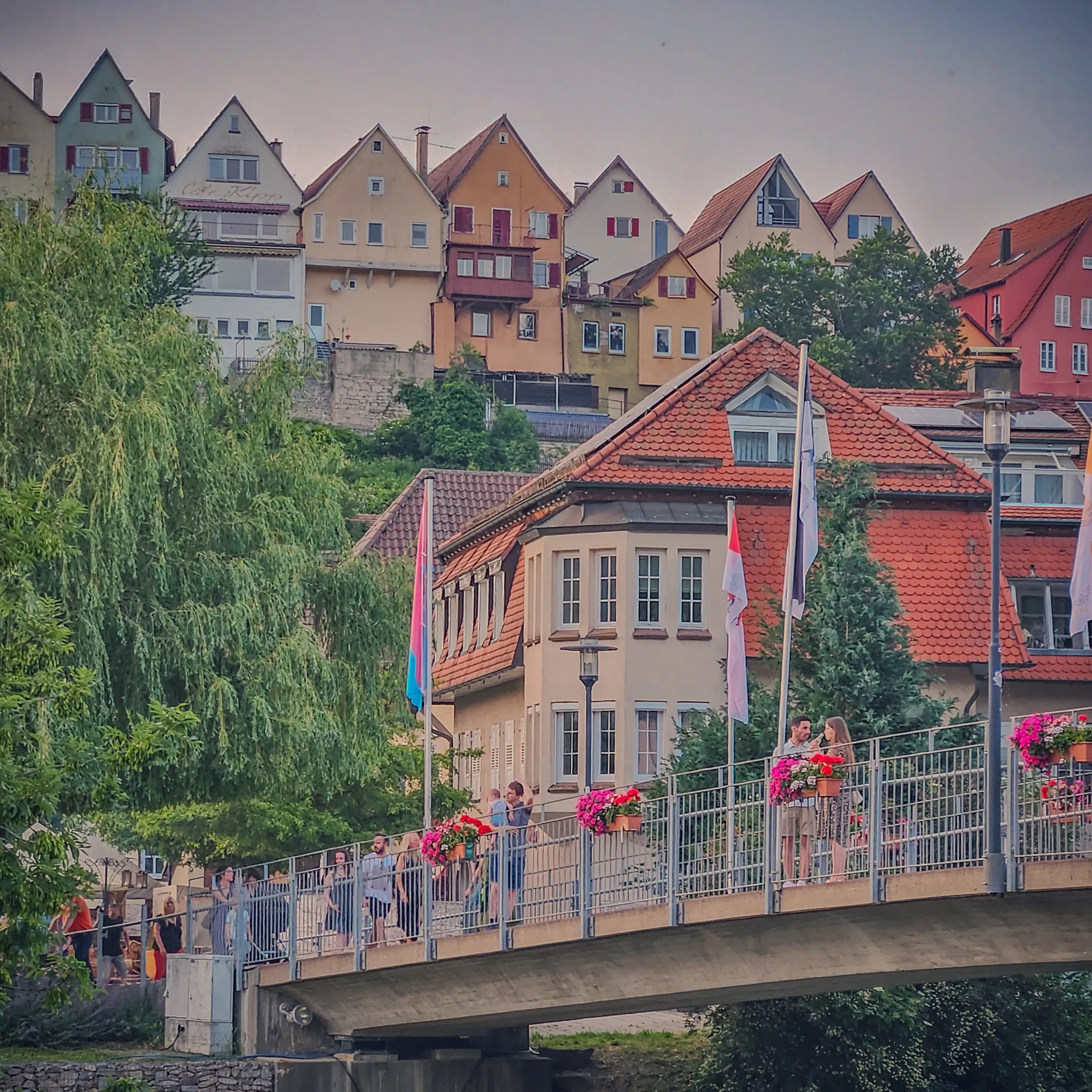 People cross a flower-decorated pedestrian bridge in Horb am Neckar, with hillside houses stacked behind them.