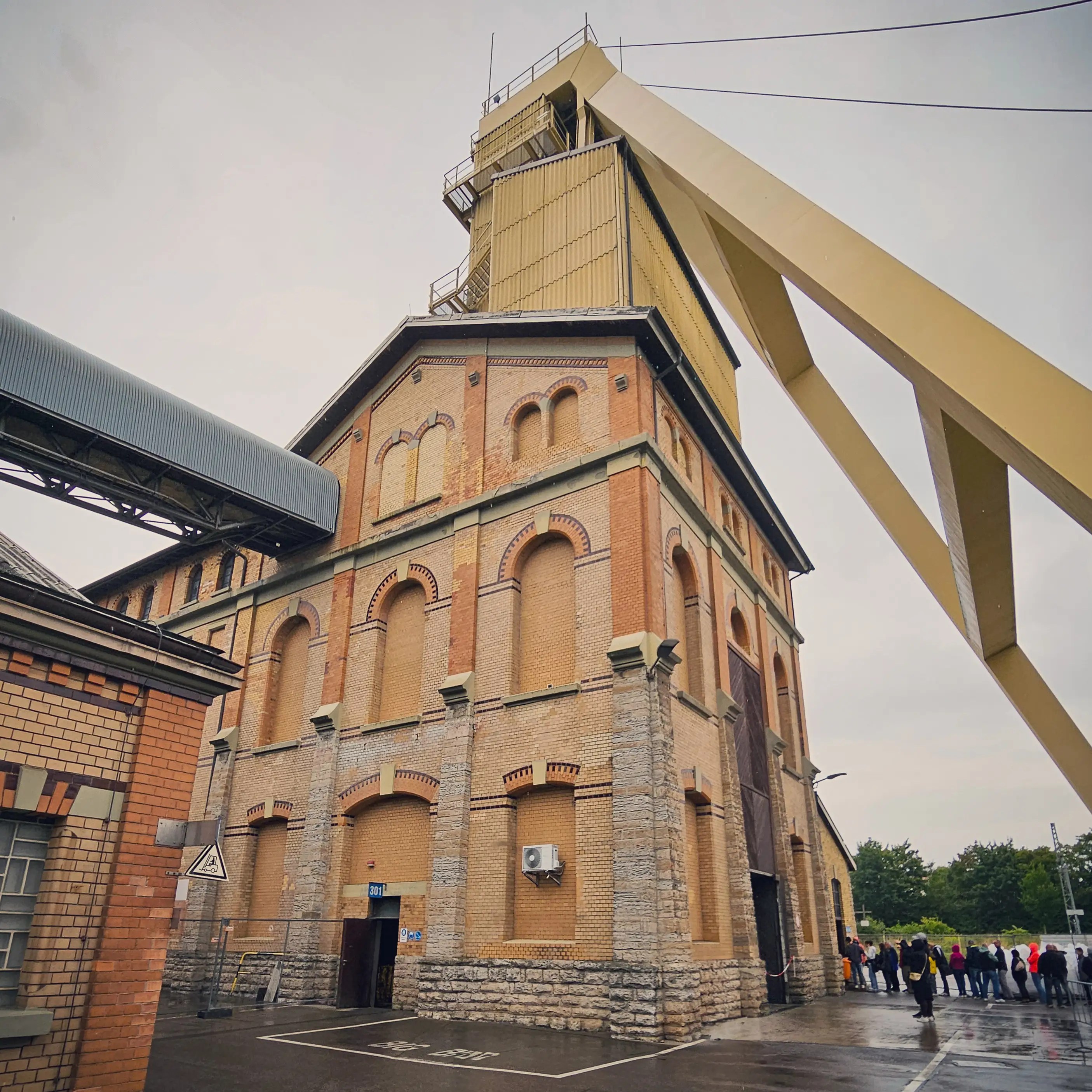 Side angle of the brick mine shaft building with its tall steel headframe and visitors gathering outside.