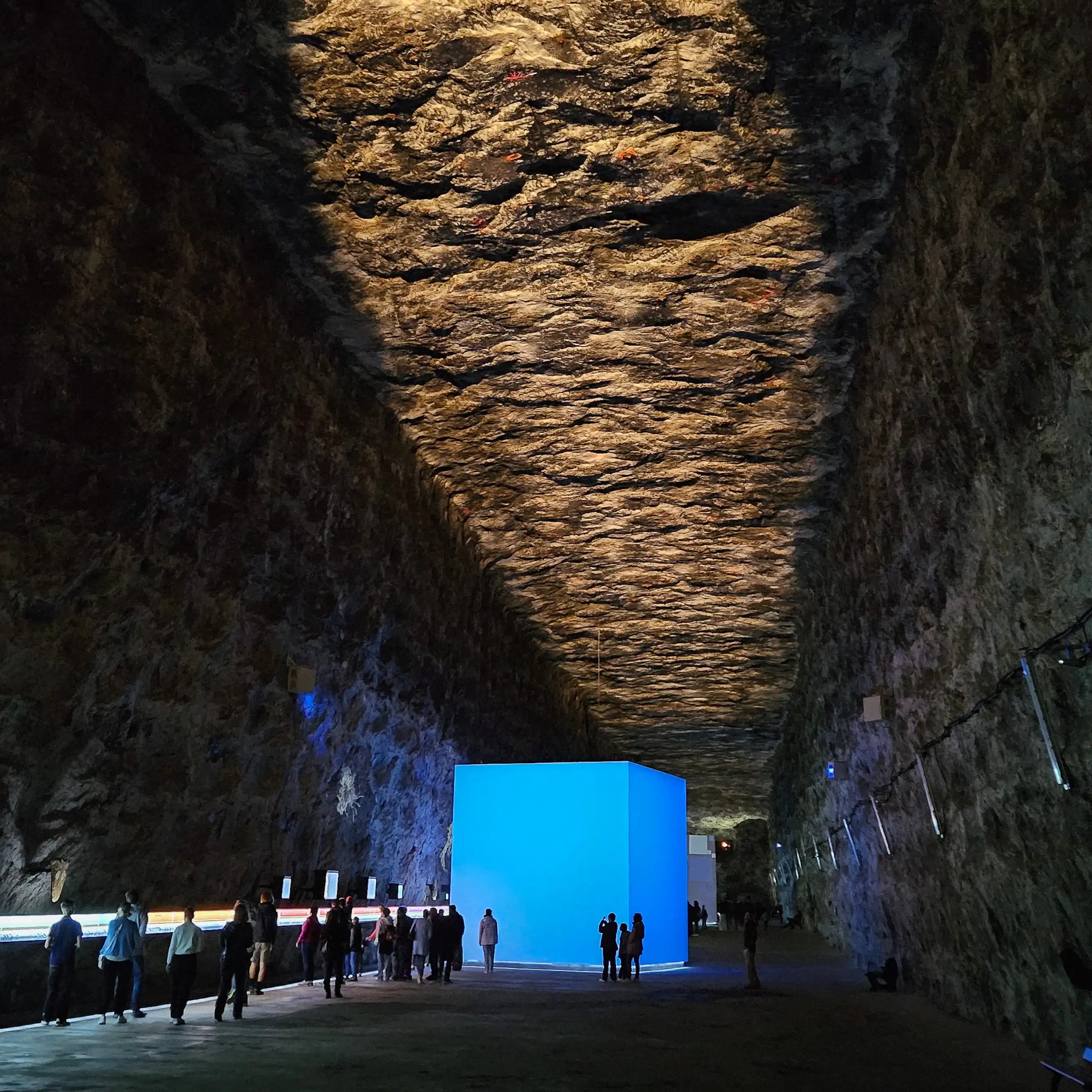 Large underground chamber with illuminated blue cube installation and visitors exploring exhibits along the walls.