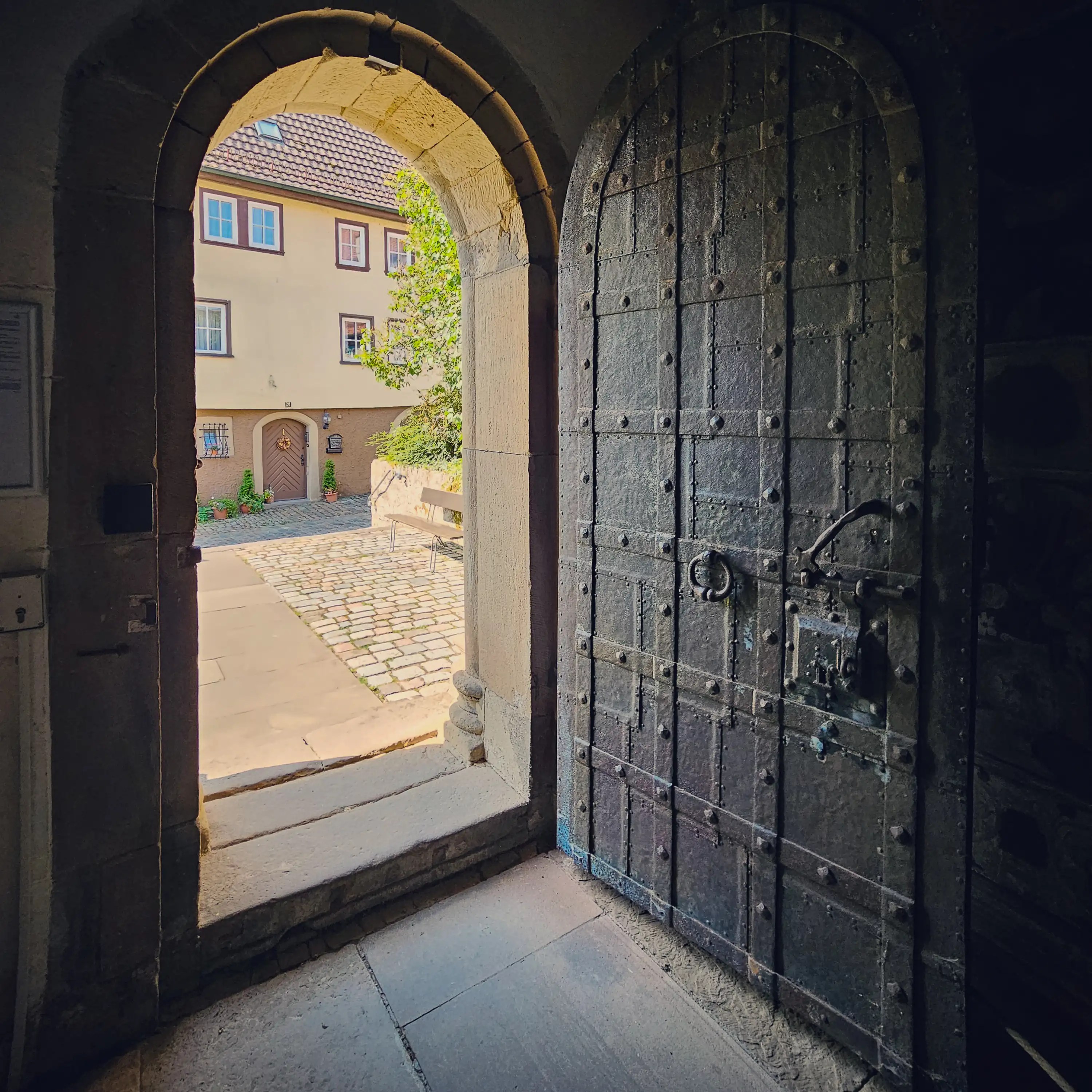 View from inside a medieval stone arch with a heavy iron-studded wooden door standing open, looking onto a sunny cobbled courtyard and a house outside.