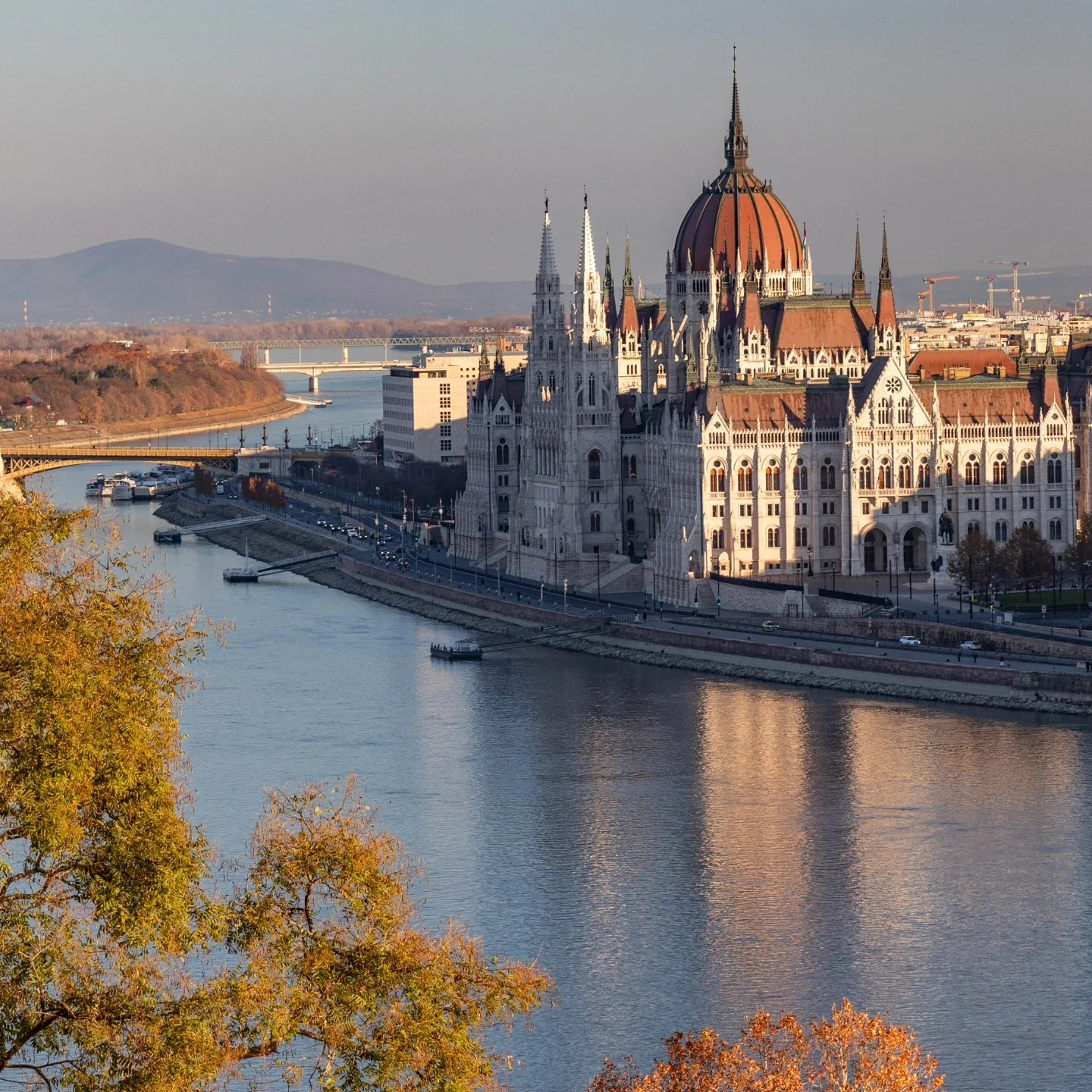 View of the Hungarian Parliament Building from across the Danube River, with soft golden sunlight reflecting off the water and autumn trees in the foreground.
