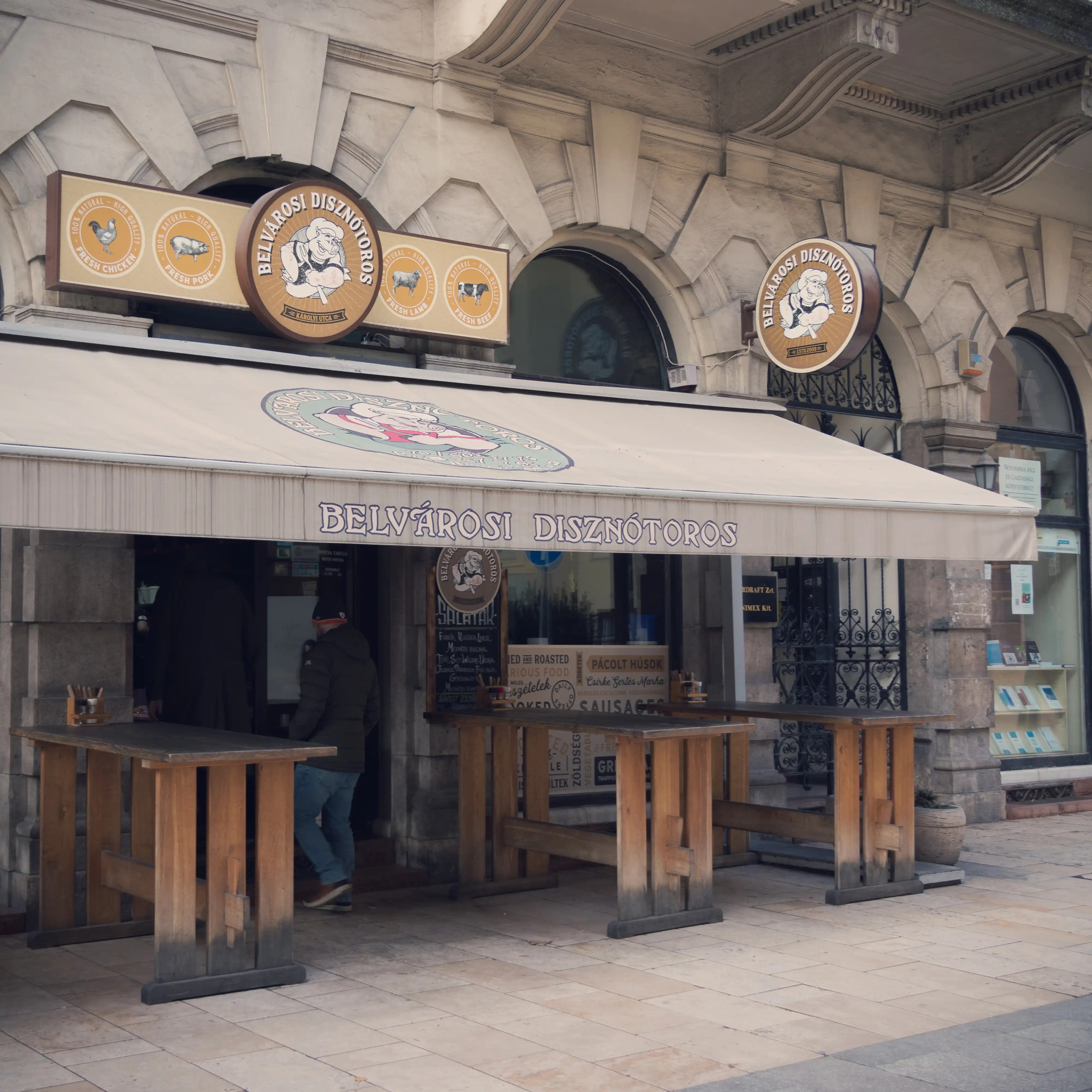 Exterior of Belvárosi Disznótoros, a Hungarian street food spot with a pig logo and wooden standing tables.