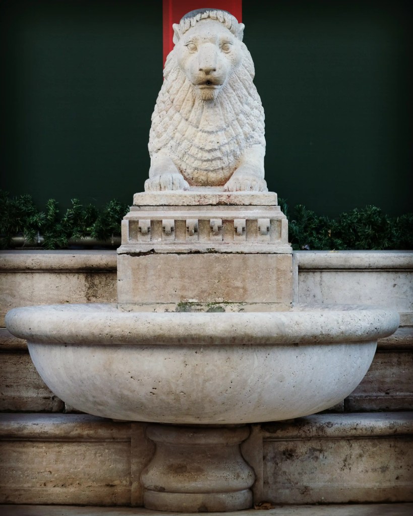 A stone lion-head fountain with a wide basin set against green and red holiday backdrops in Budapest.