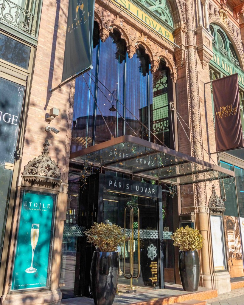 Exterior of the Párisi Udvar Hotel in Budapest, featuring arched windows, decorative stonework, and a modern glass entrance with festive holiday lights.