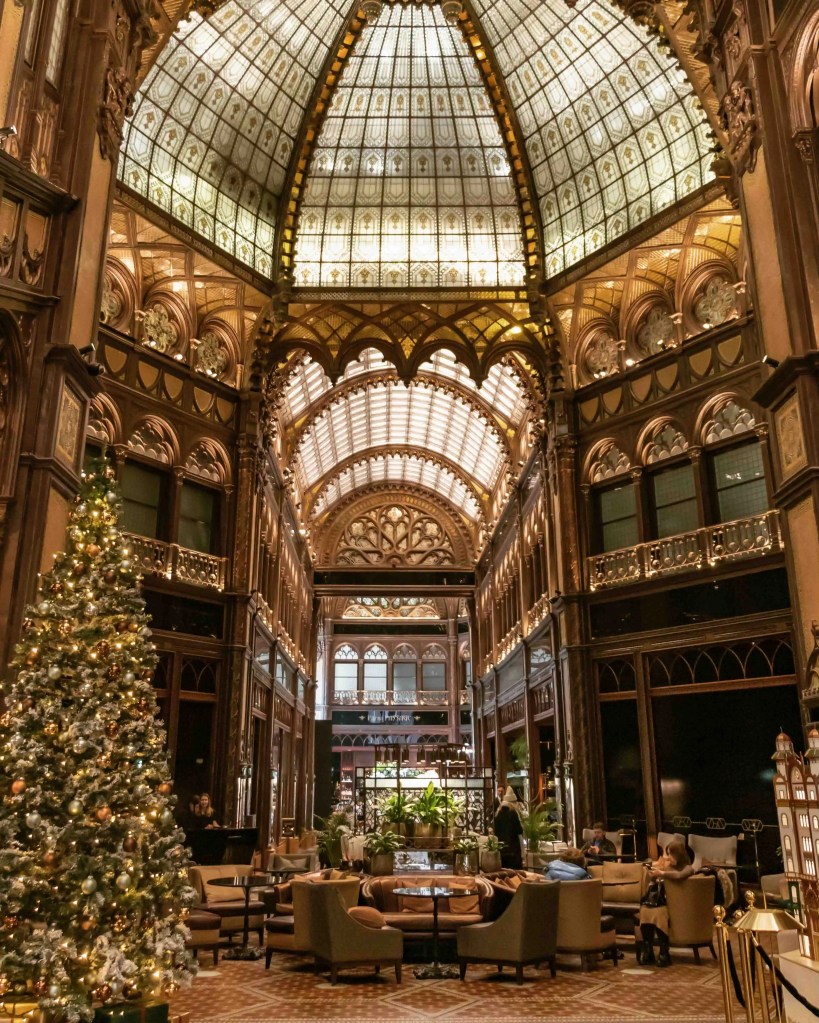 The ornate glass-domed interior of the Párisi Udvar Hotel in Budapest, with festive Christmas decorations, plush seating, and intricate architectural details.