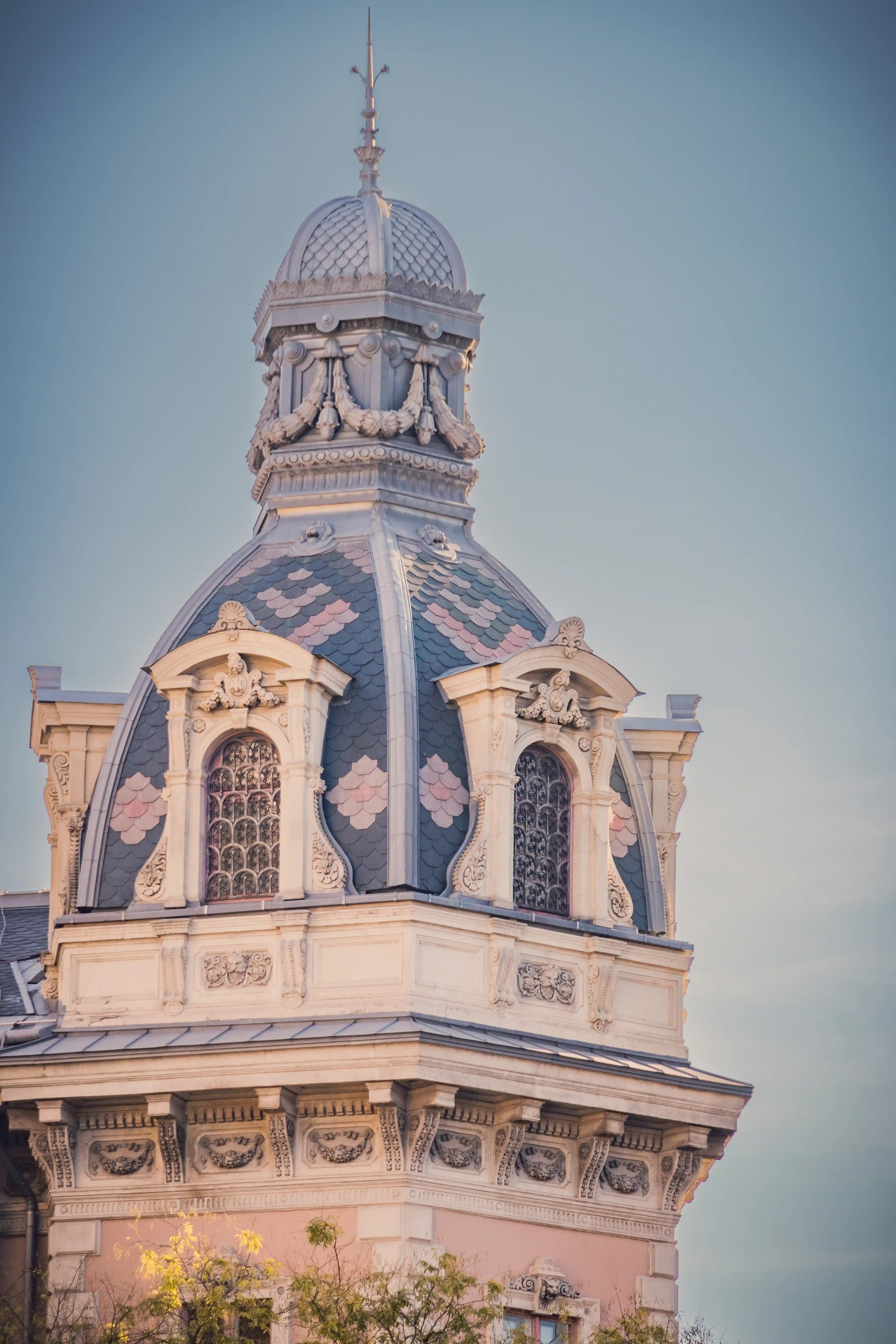 Decorative rooftop dome with pink and blue tiles and ornate windows, photographed in Budapest.