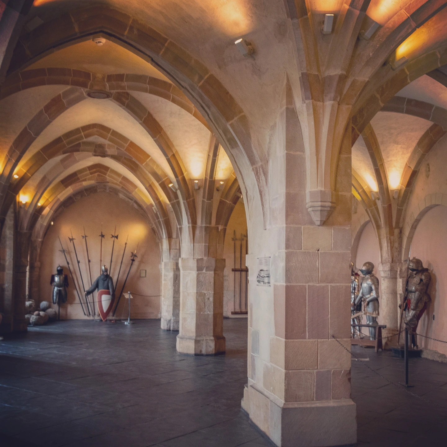 Stone vaulted armory in Vianden Castle displaying suits of armor and weapons.