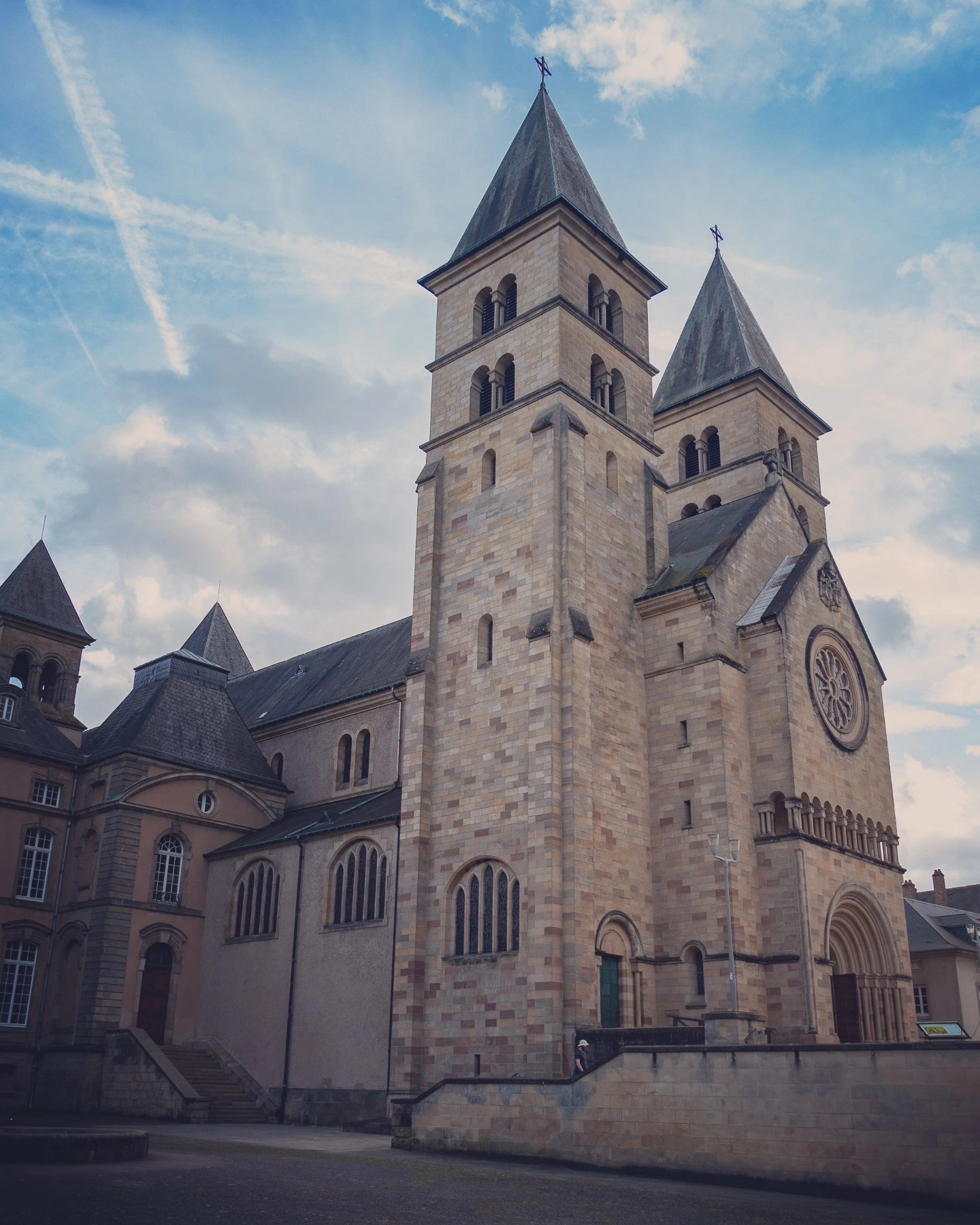 Exterior of the Basilica of St. Willibrord in Echternach, Luxembourg, showing its Romanesque twin towers and rose window under a blue sky.