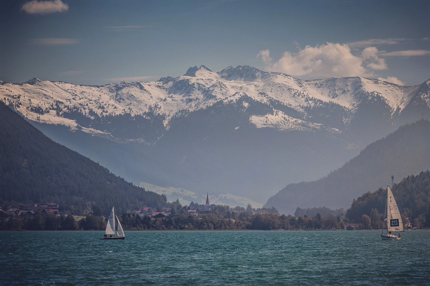 Sailboats on emerald Achensee with snow-dusted mountains and soft clouds in the distance.