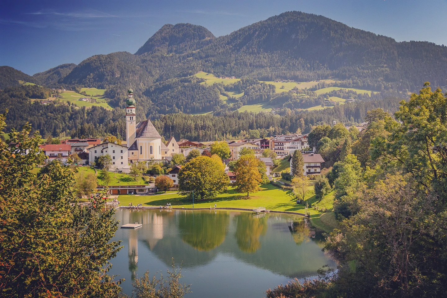 Reith im Alpbachtal village and church reflected in a small lake with forested mountains behind.
