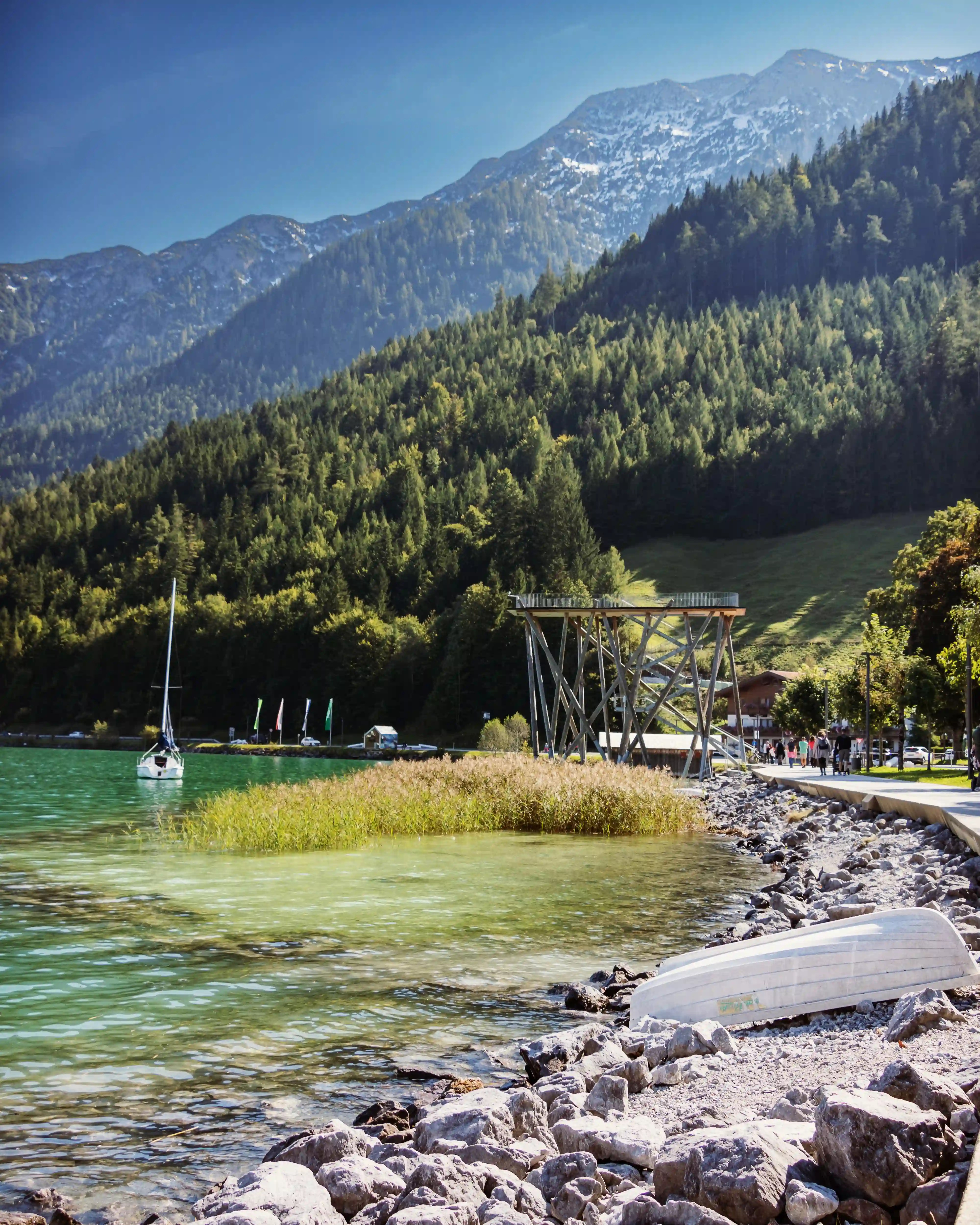 Clear green water of Lake Achensee along a rocky shore with a sailboat and pine-covered mountains in the background.
