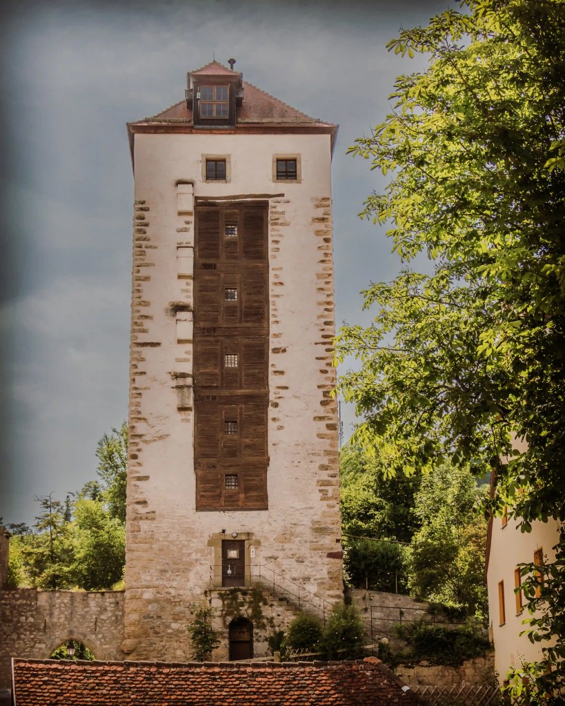 A tall medieval stone tower in Horb am Neckar rises above trees and historic rooftops, partially lit by sunlight.