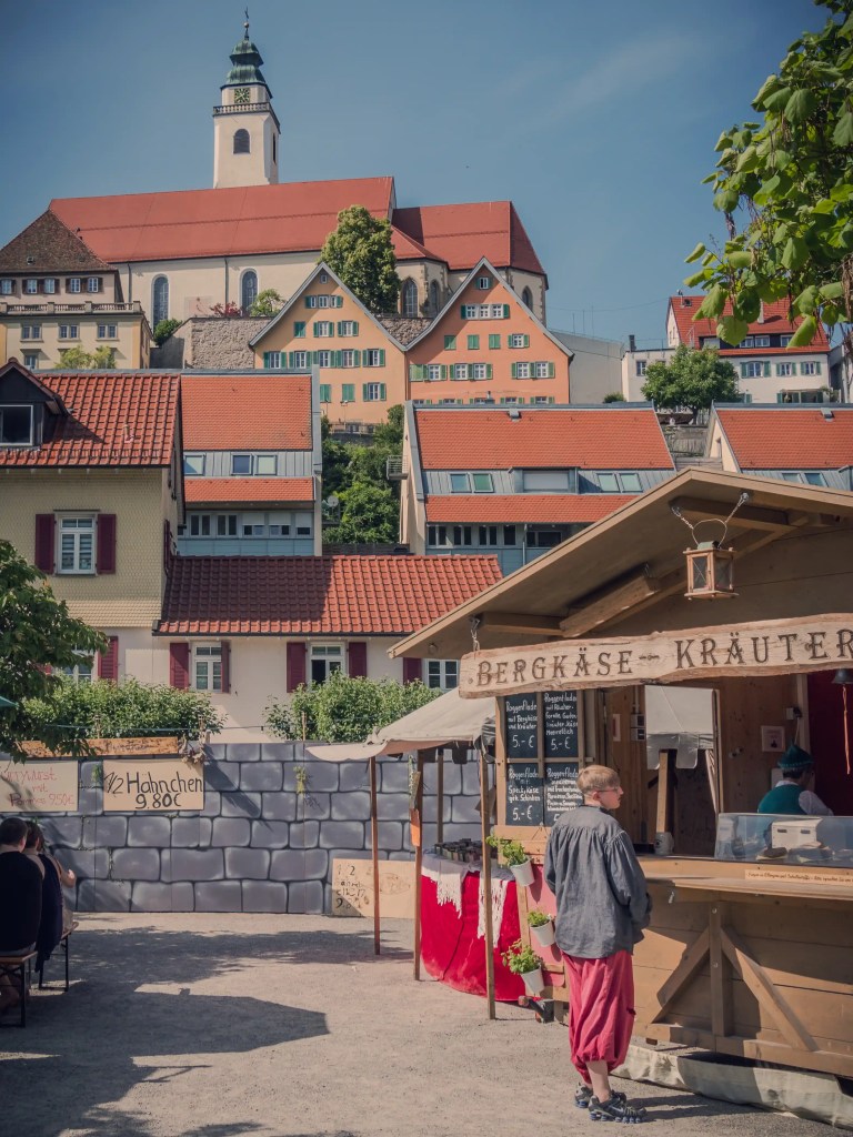 A small medieval-style food stall with a handwritten sign advertising “½ Hähnchen” (half chicken) and a church and colorful hillside homes rising behind.