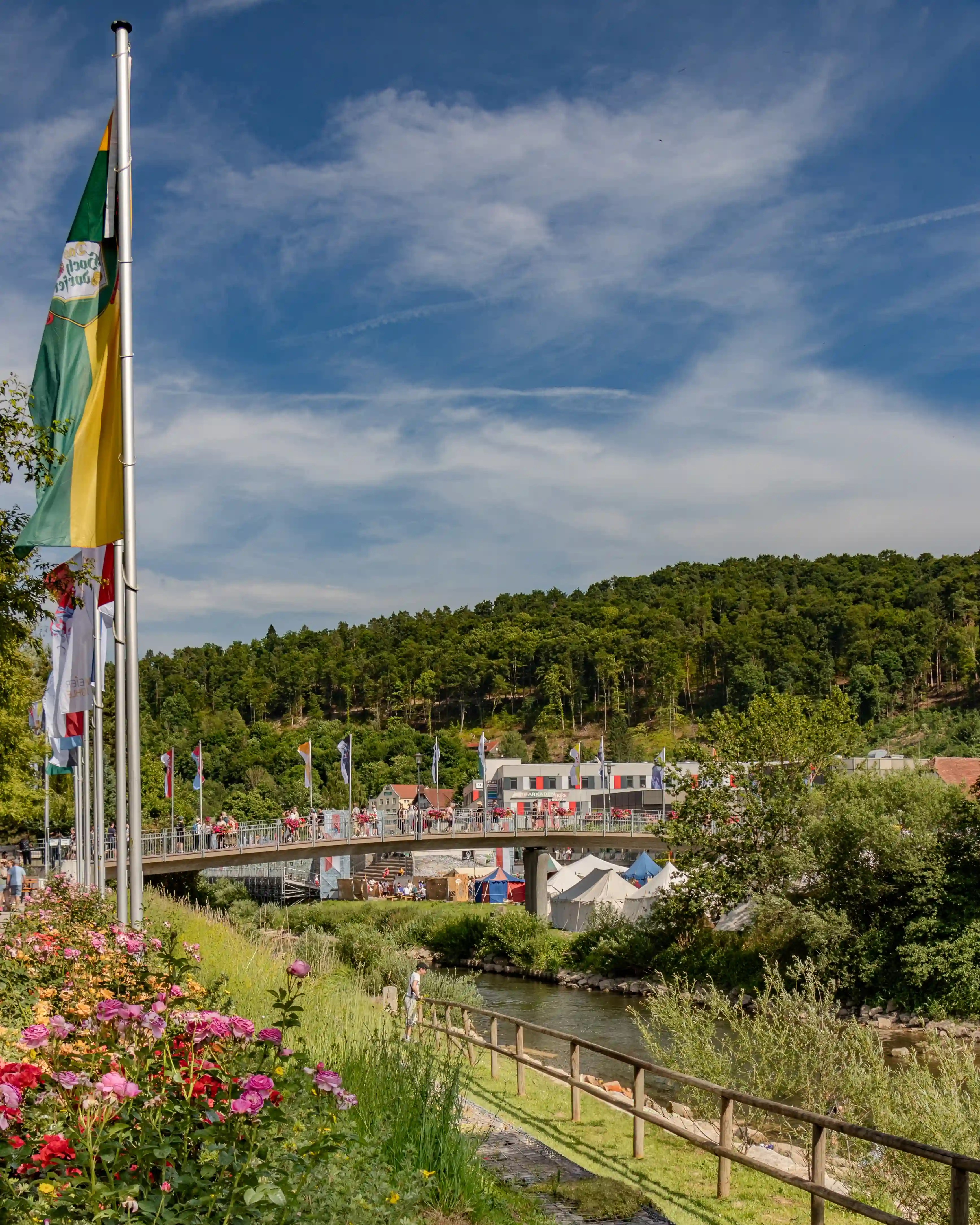 View across a bridge lined with flags and people, overlooking a tent village and lush green hillside in the background.