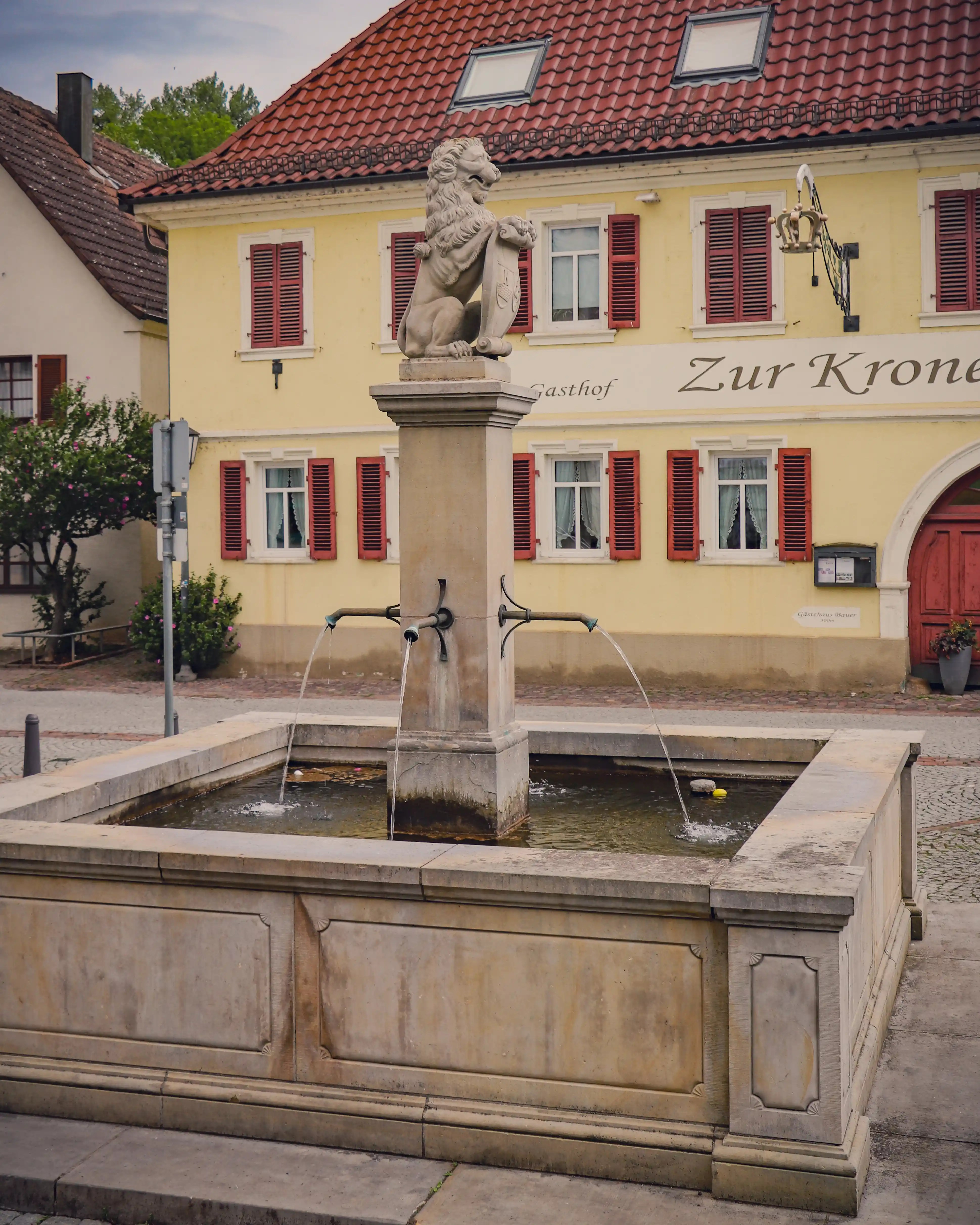 Stone fountain with a lion statue holding a scroll in Bad Friedrichshall’s town square, with the Gasthof Zur Krone in the background.