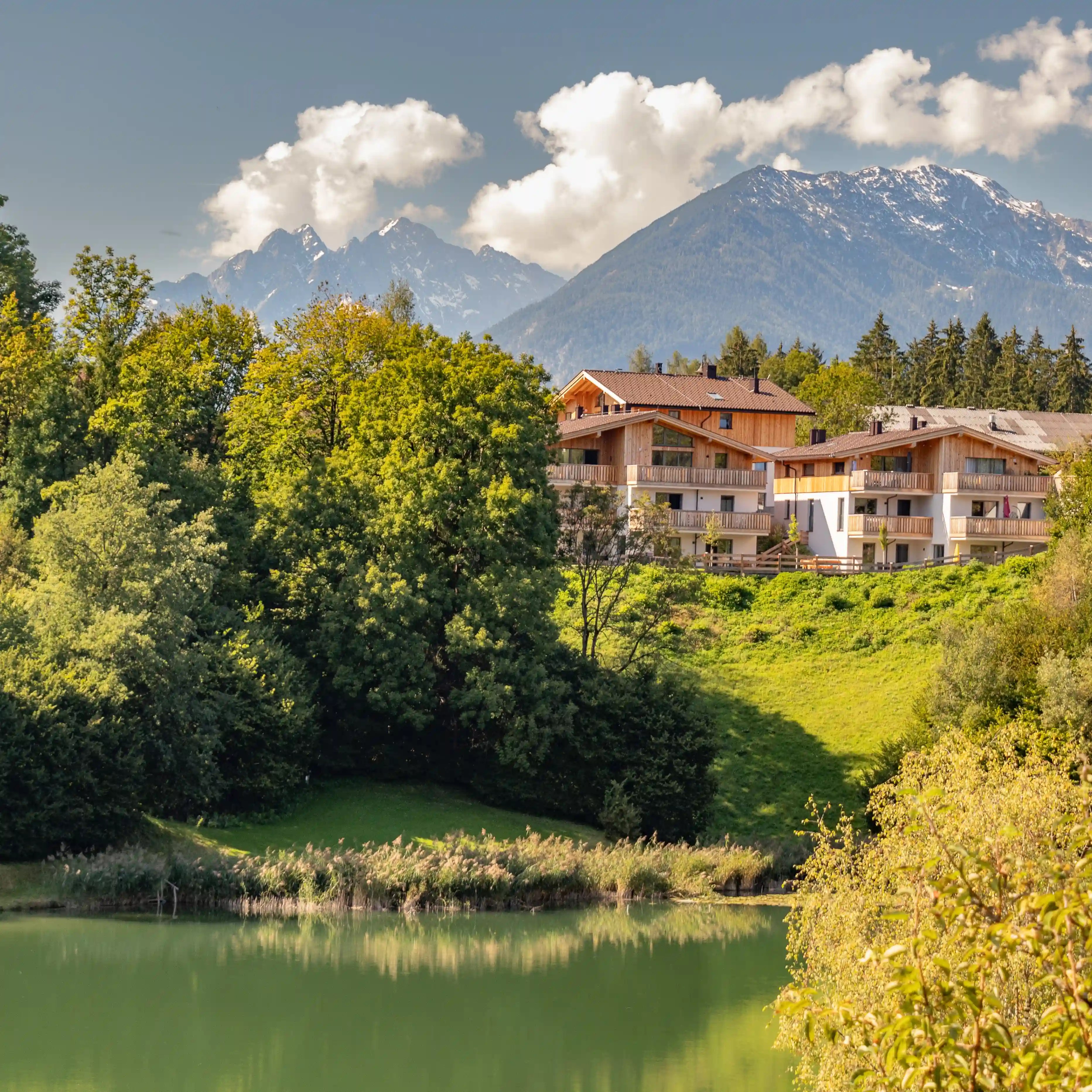 Chalet-style apartments on a green hillside reflected in a calm pond with distant mountains.