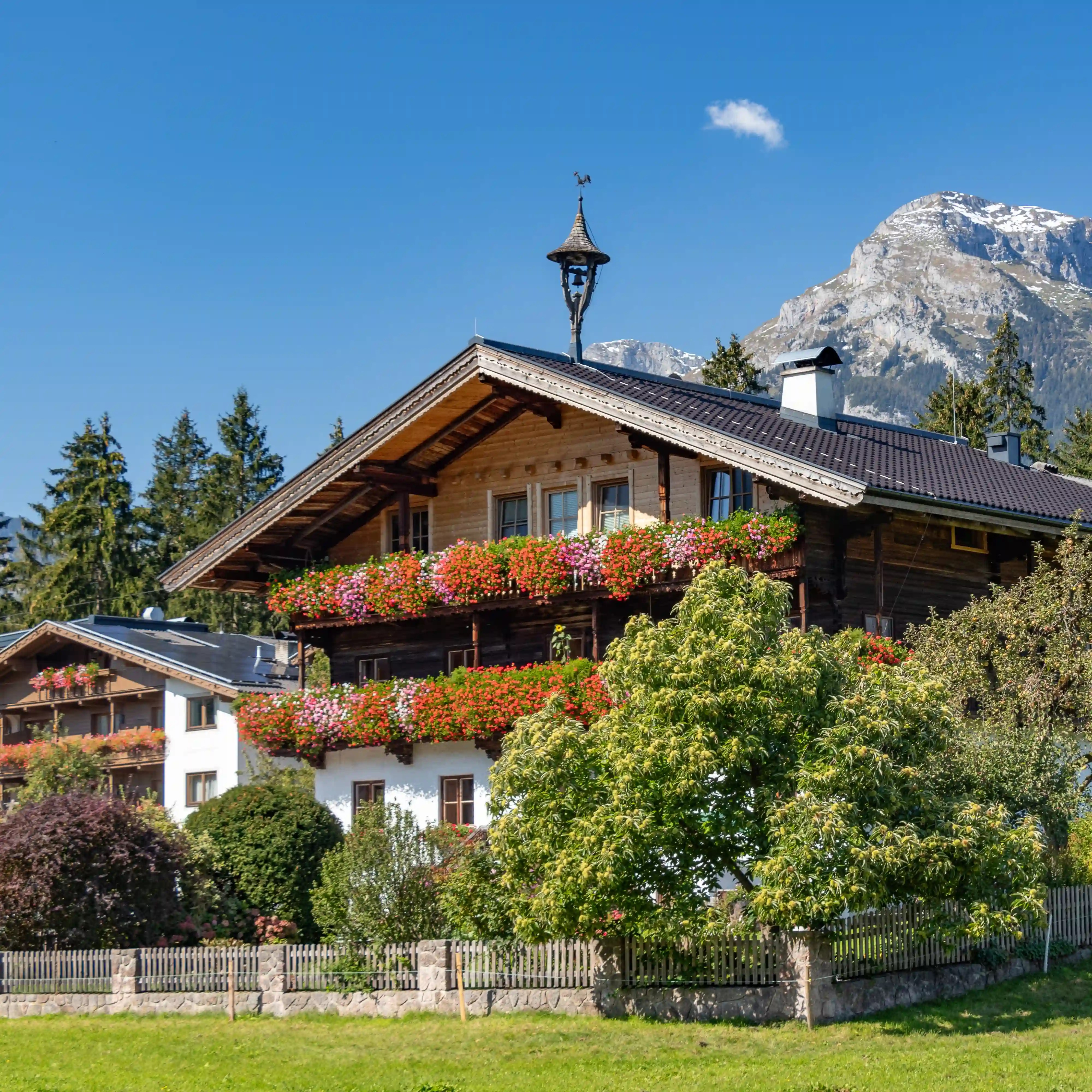 Traditional farmhouse with two tiers of flower boxes, set before trees and buses with a backdrop of rocky alpine peaks.