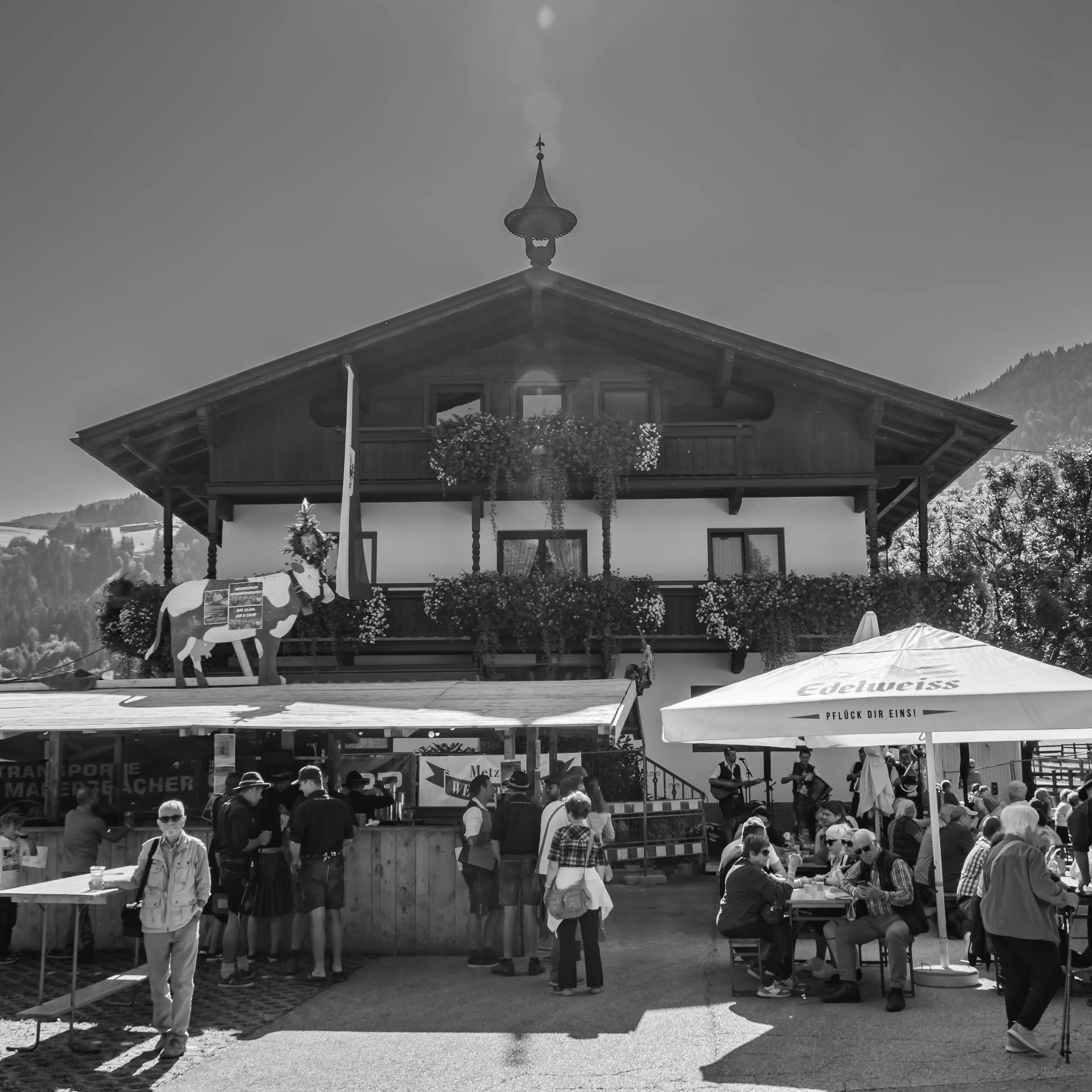 Black-and-white street scene of festival stalls and people dining in front of a gabled farmhouse.