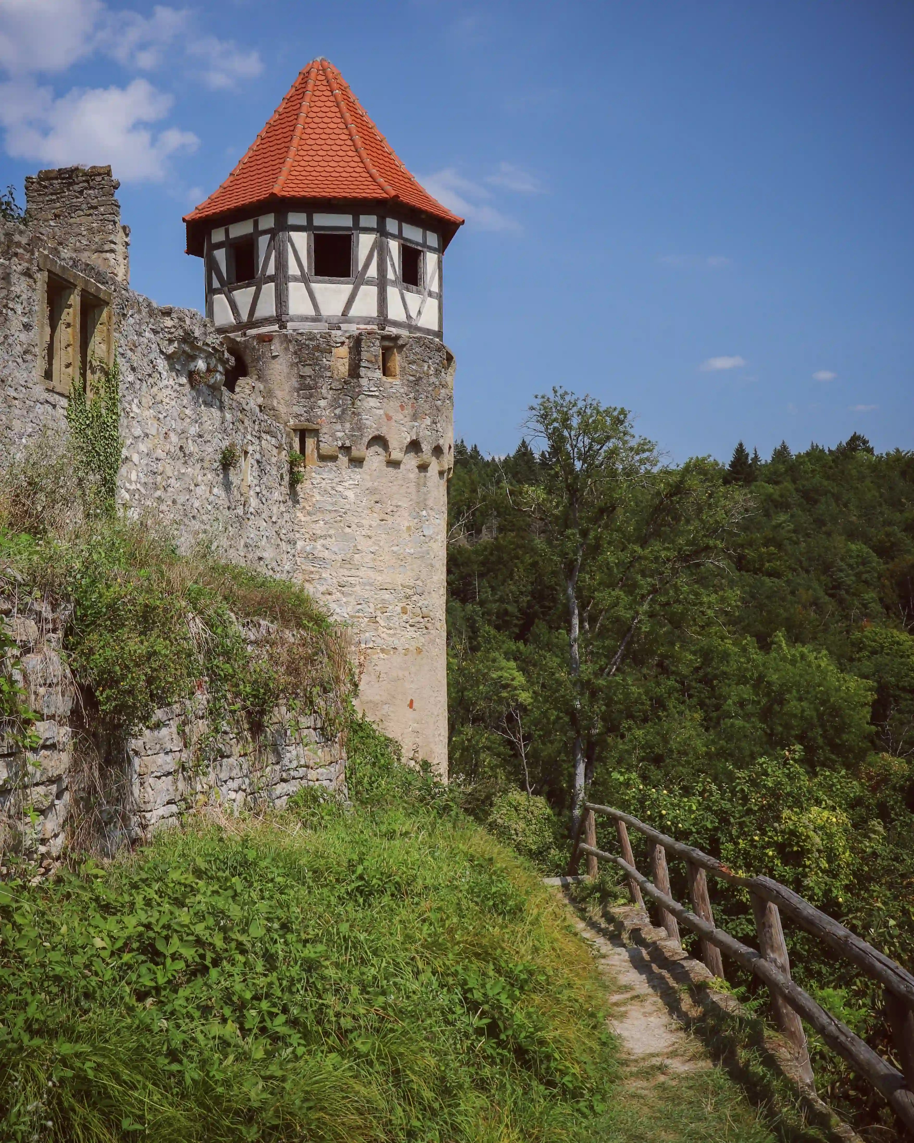 Stone ramparts and a timbered turret with crenellations beside a narrow hillside path and wooden rail.