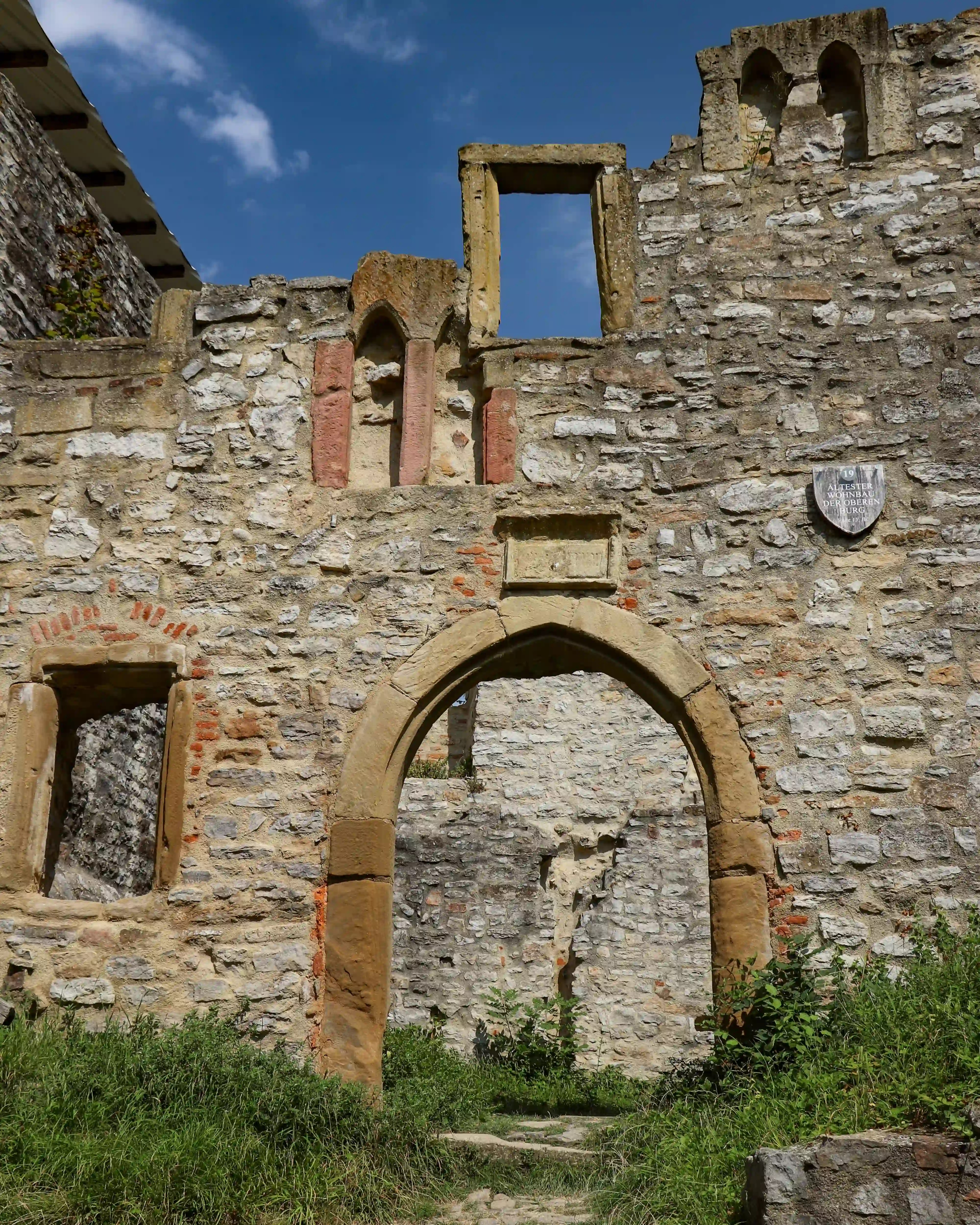 Ruined stone wall with pointed Gothic arch and fractured windows at Burg Hornberg, grasses growing below.
