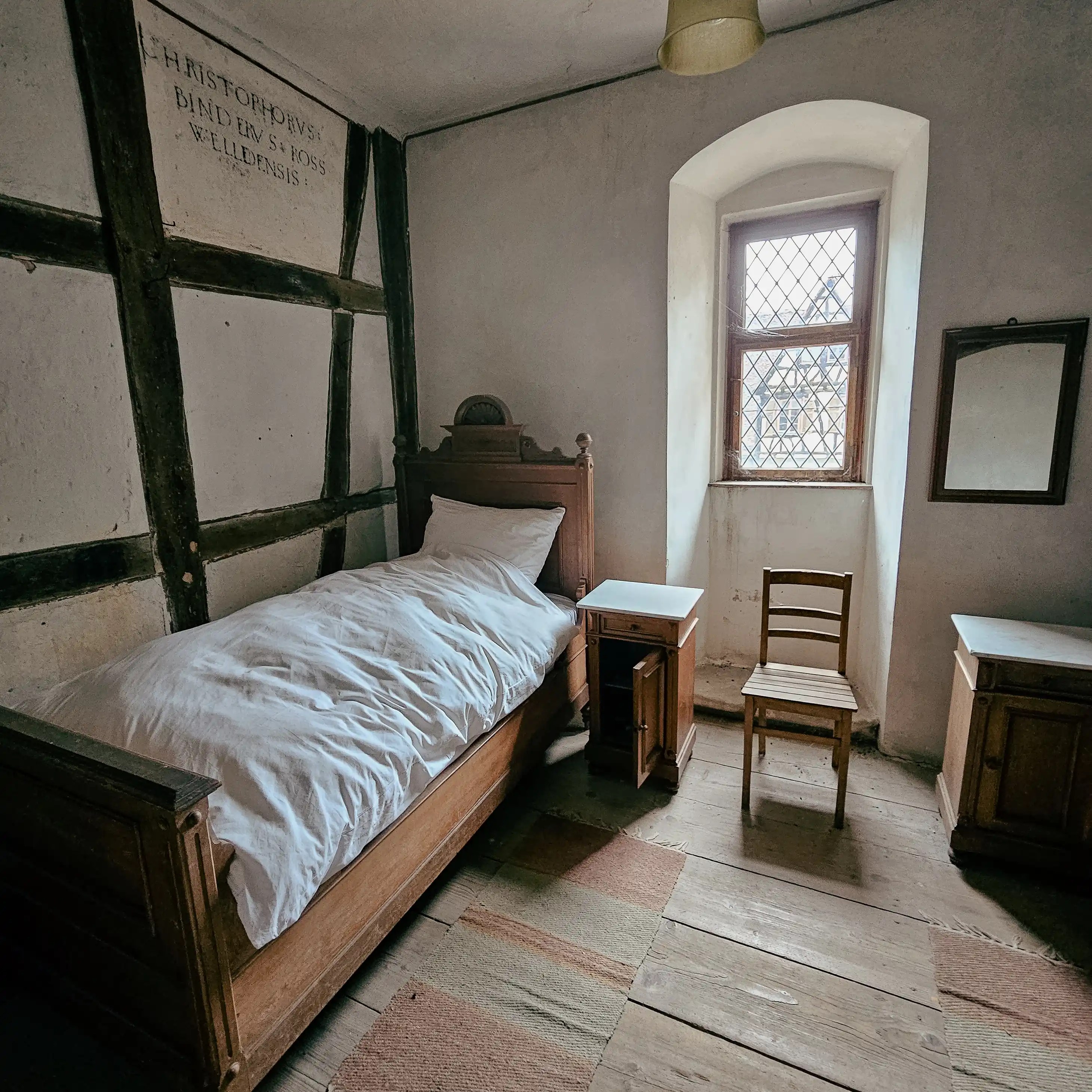 Simple monastic bedroom with wooden bed, chair, and window in Bebenhausen Monastery.