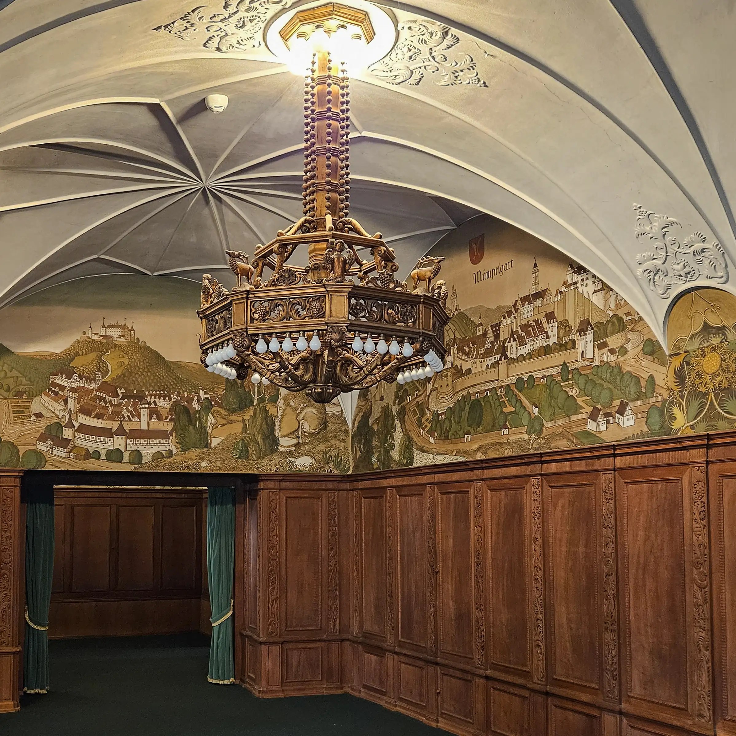 Palace room at Bebenhausen with wood-paneled walls, mural of a town, vaulted ceiling, and ornate chandelier.