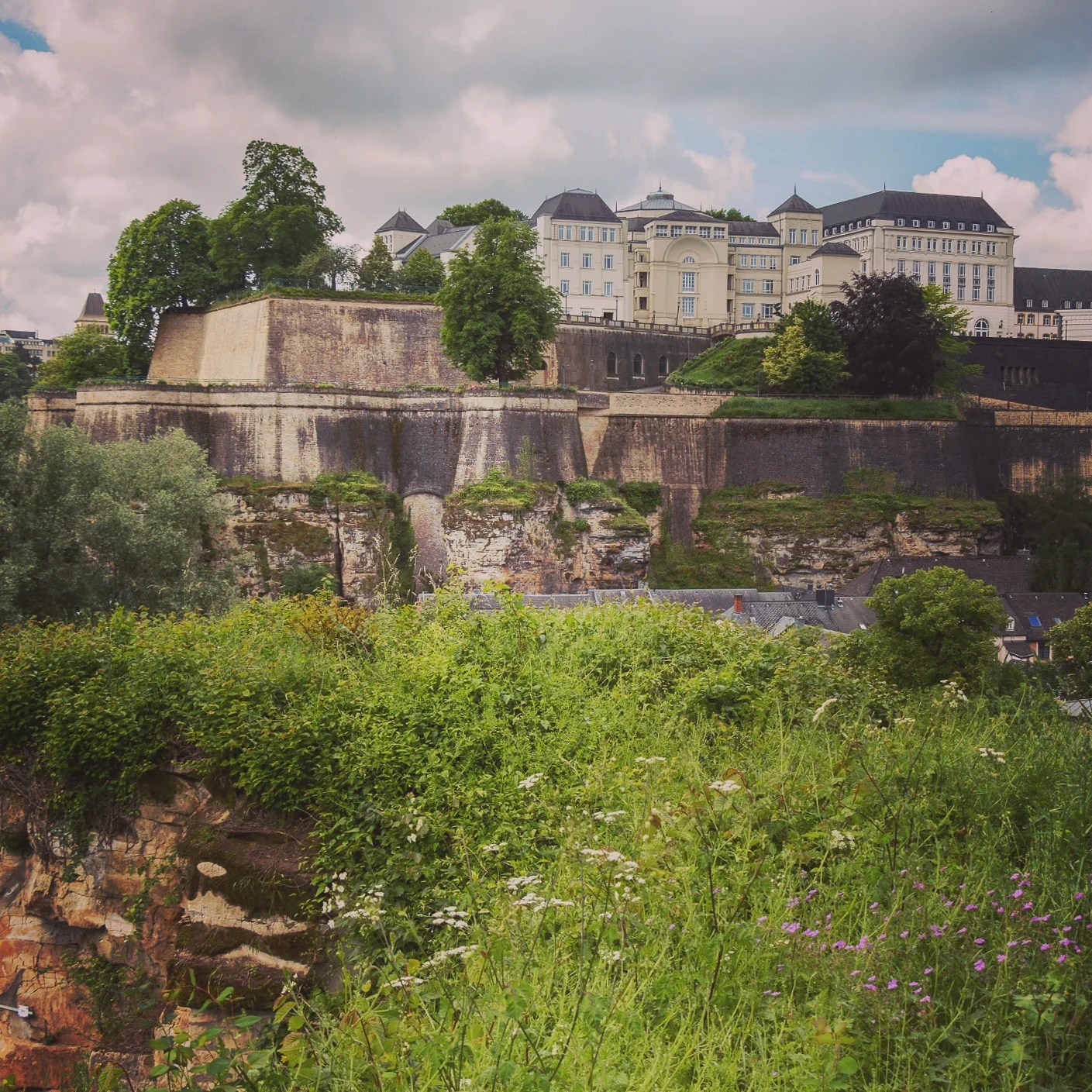 Massive stone fortress walls of Luxembourg City with greenery and historic buildings.